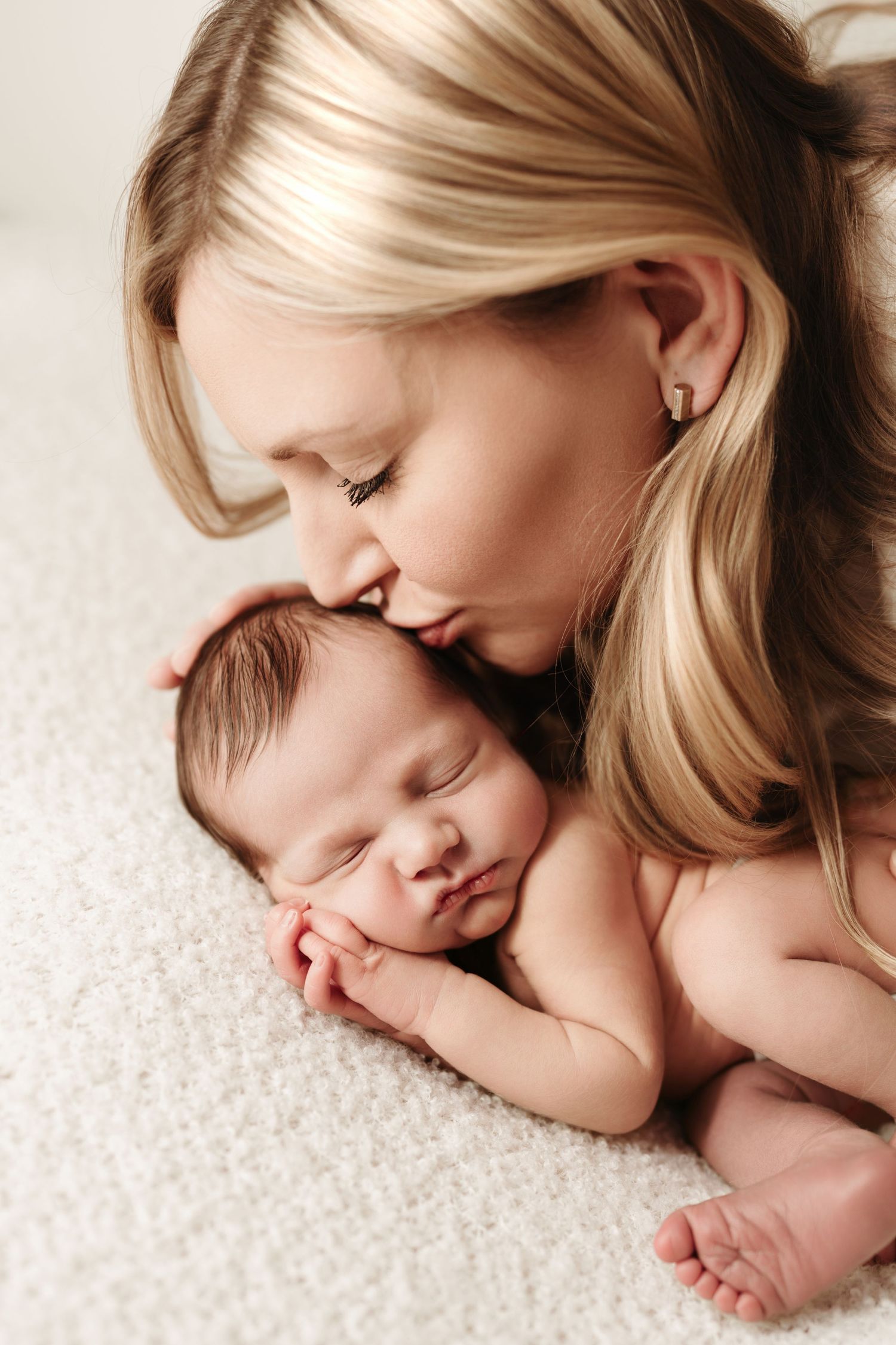 Blonde mother tenderly kissing her sleeping newborn baby on white fluffy blanket.