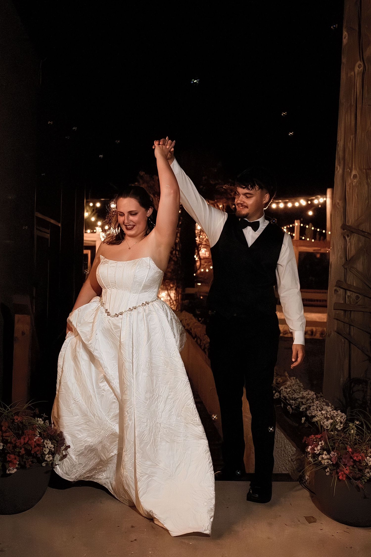 Bride and groom make grand entrance with raised hands under string lights at rustic barn wedding venue.