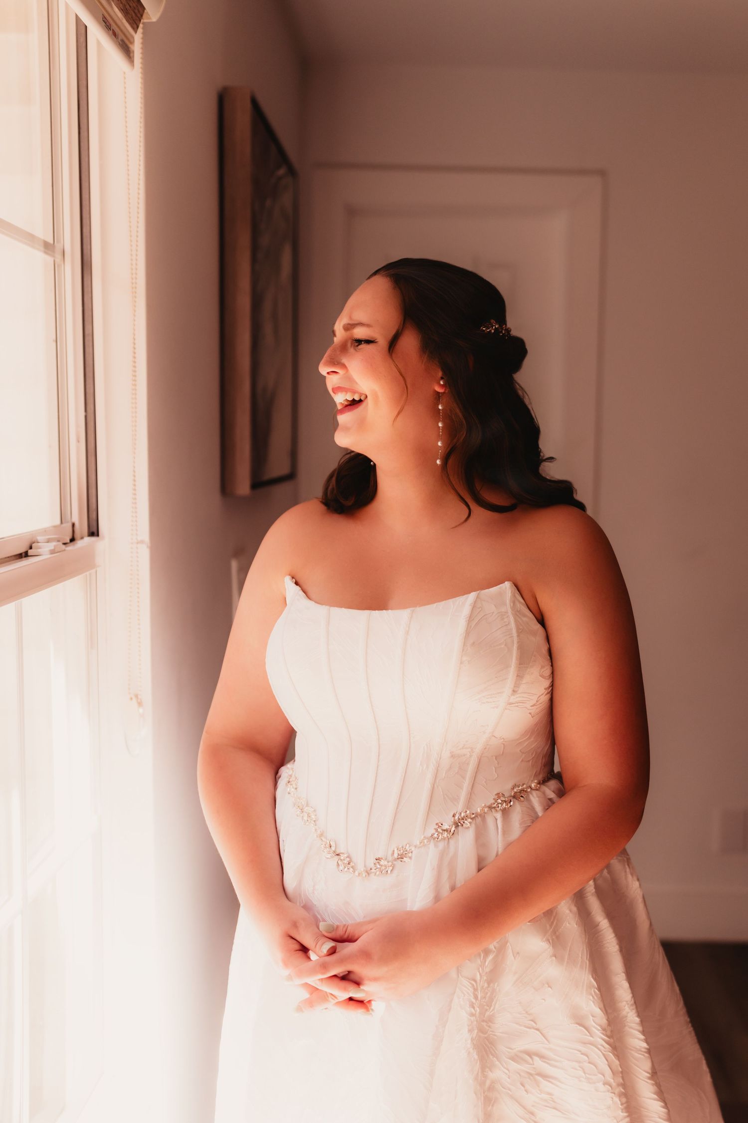 Radiant bride in strapless white gown with crystal belt laughs joyfully while standing near sunlit window.