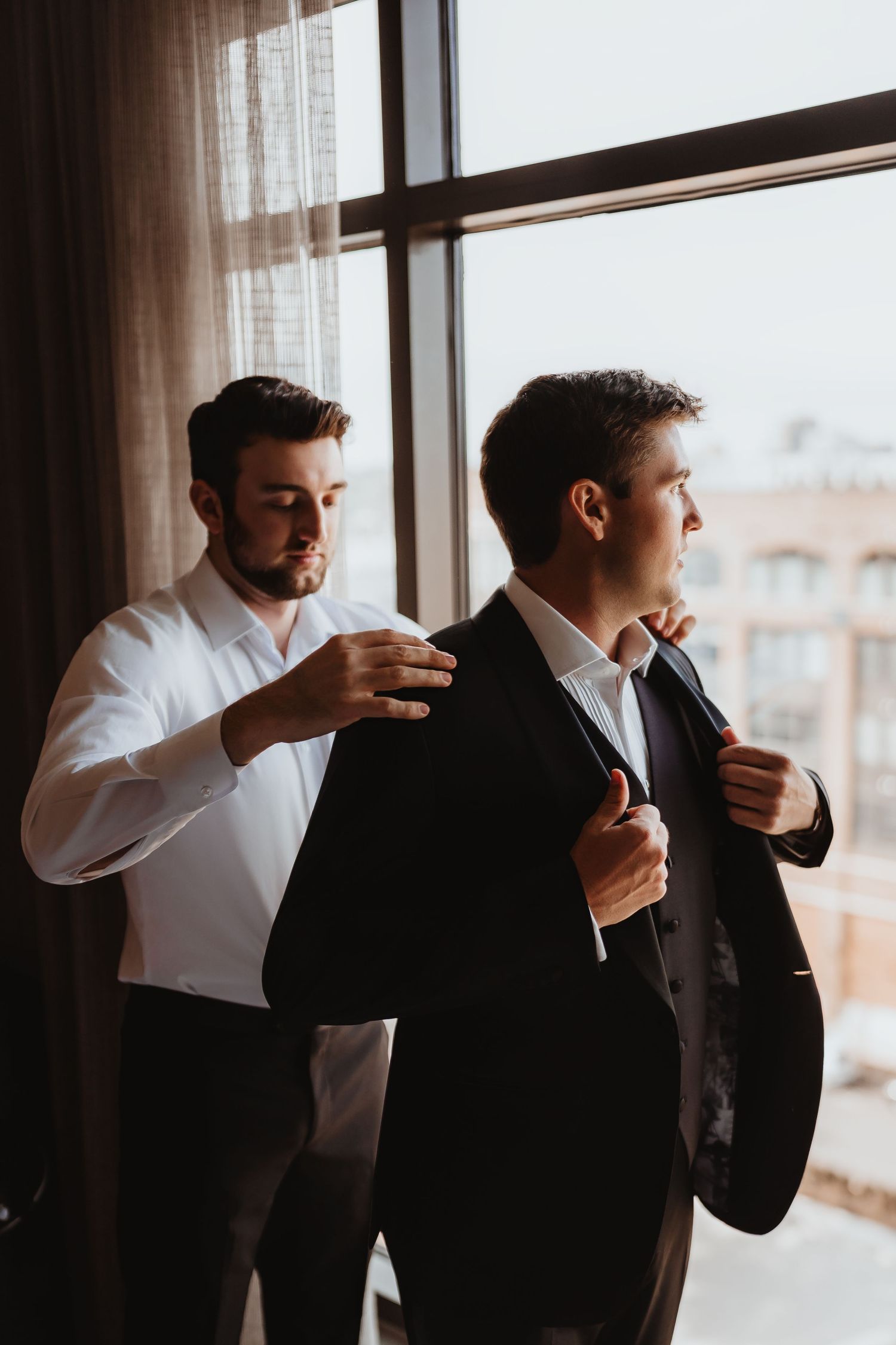 Best man helping groom button dark suit jacket near large window with city view behind them.