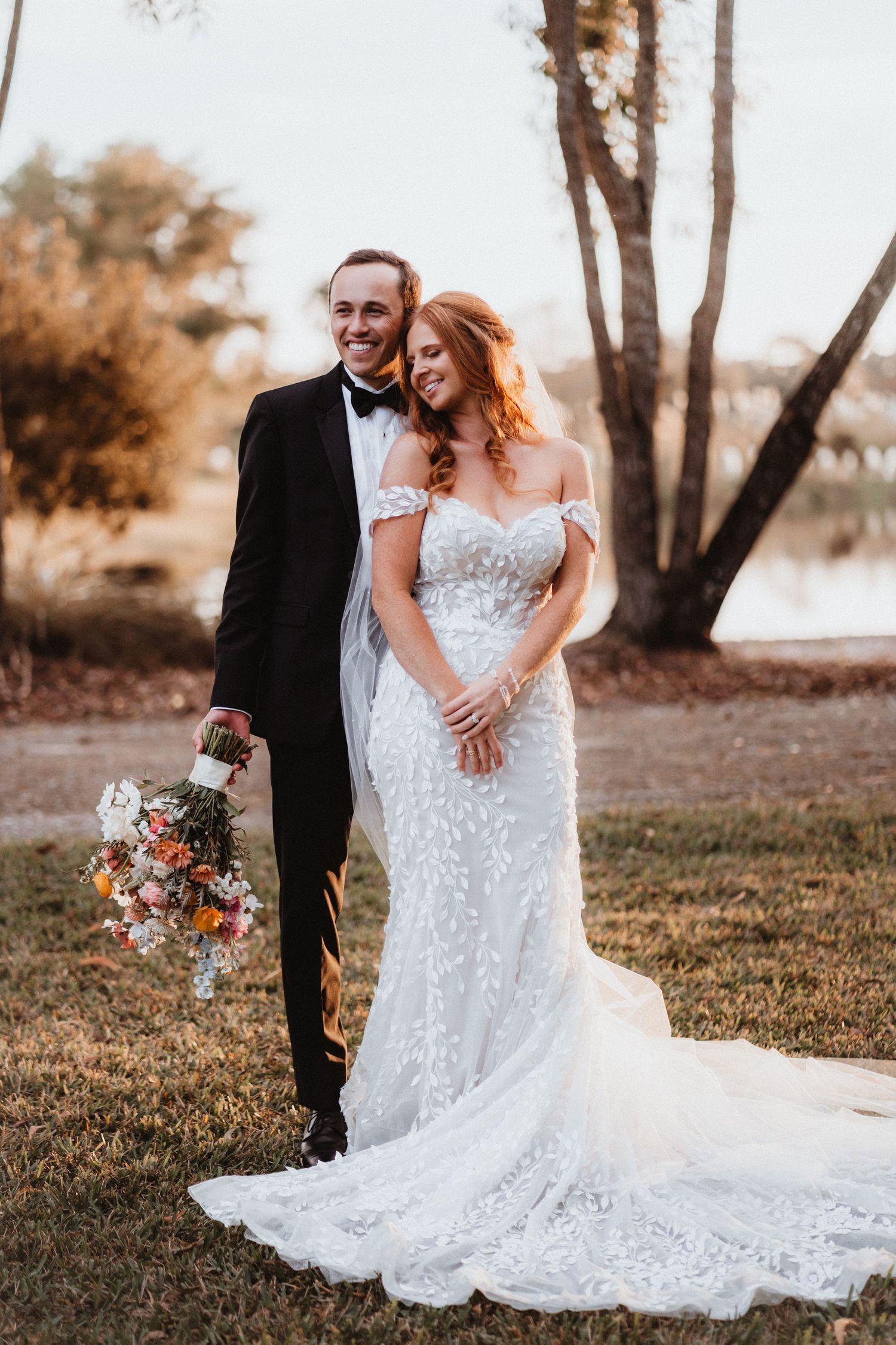 Smiling bride and groom pose outdoors at golden hour, groom holding colorful floral bouquet in black tuxedo.
