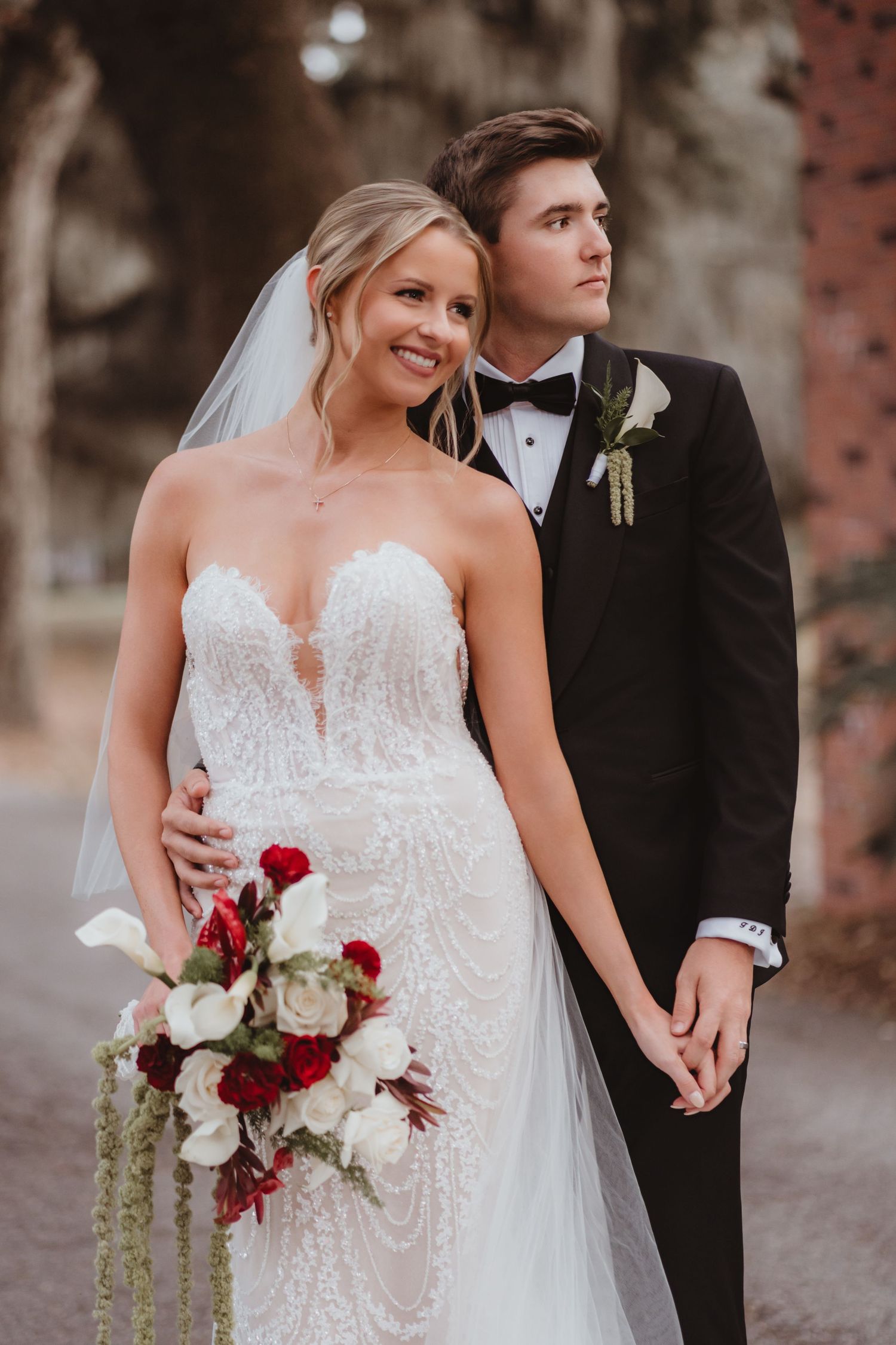 Smiling bride in strapless lace gown holds red and white bouquet as groom in tuxedo embraces her.