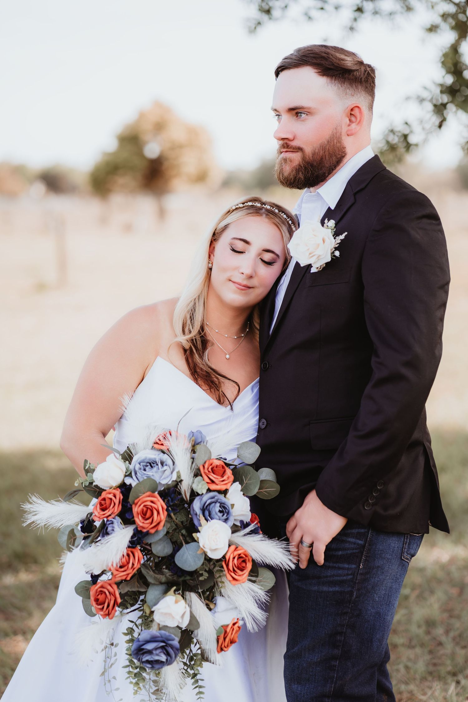 Bride resting head on groom's chest in black suit holding colorful bouquet in outdoor field setting.