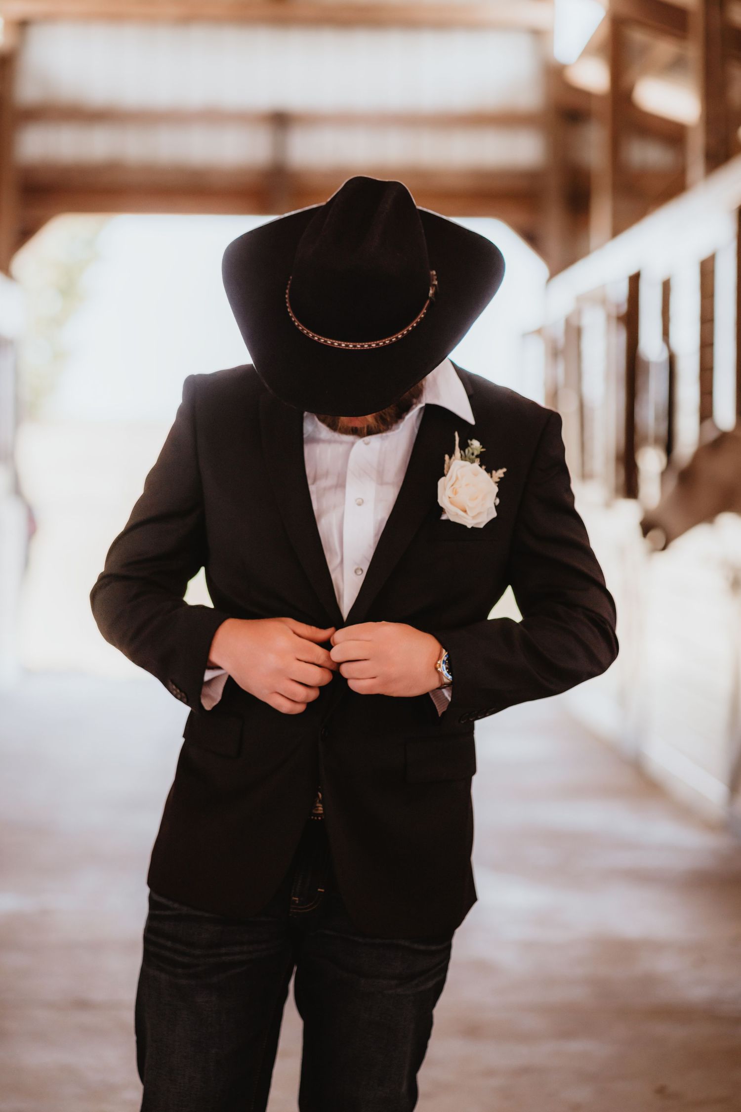 Dapper western groom in black cowboy hat buttons black blazer with white rose boutonniere in barn.
