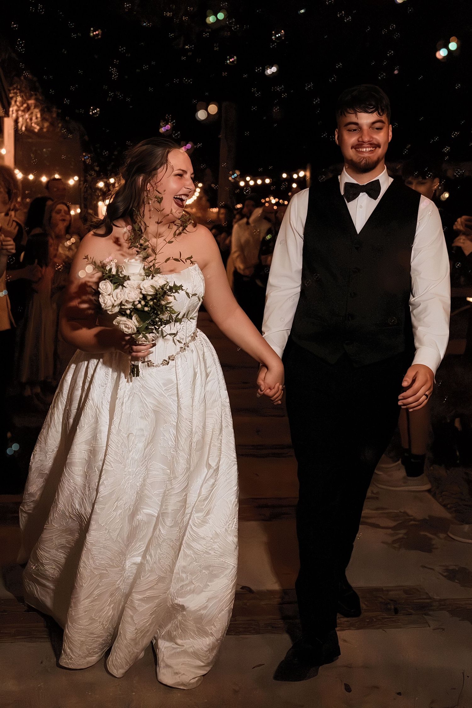 Smiling newlyweds walk hand in hand during outdoor evening wedding send-off with guests and string lights.