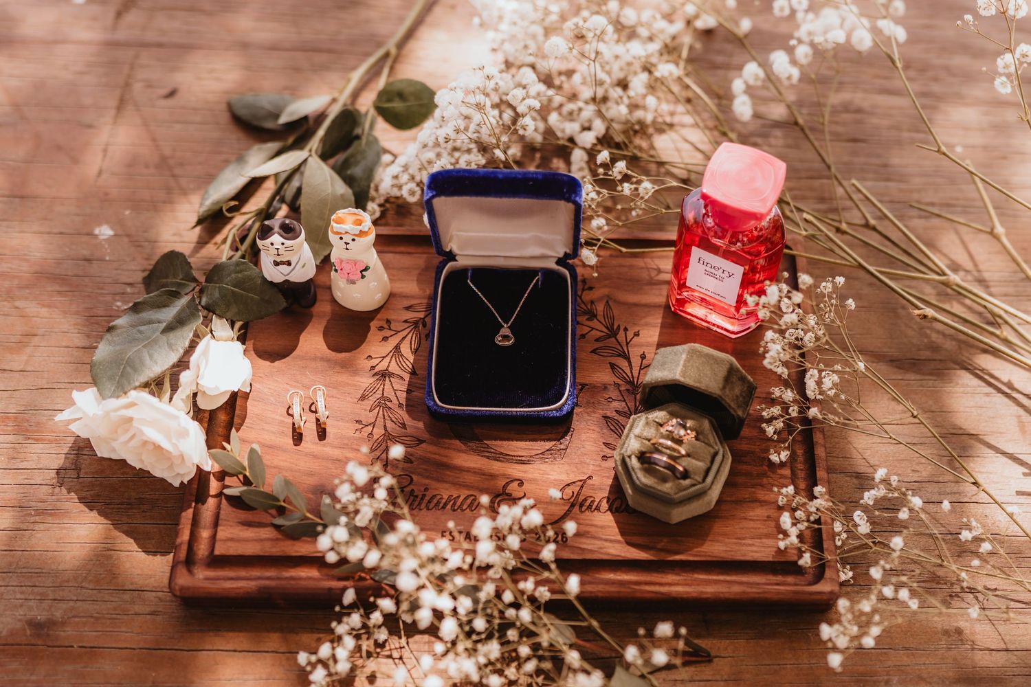 Elegant wedding flat lay with gold necklace in blue velvet box, perfume, flowers, and jewelry on wooden tray.