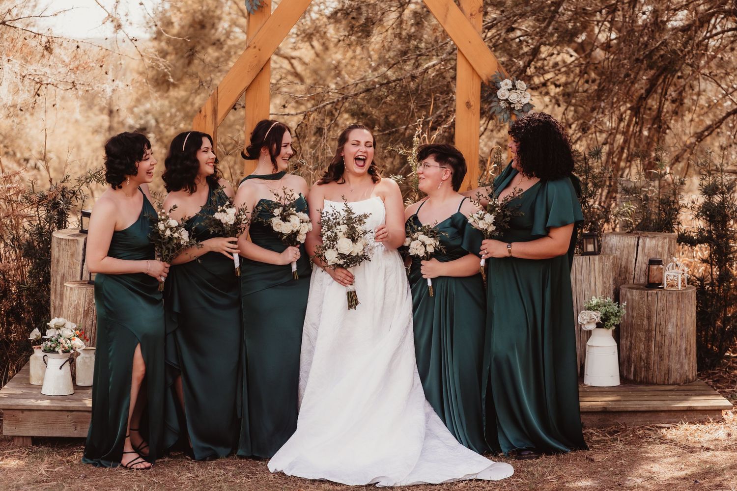 Bride in white gown laughing with four bridesmaids in emerald green dresses at outdoor wedding.
