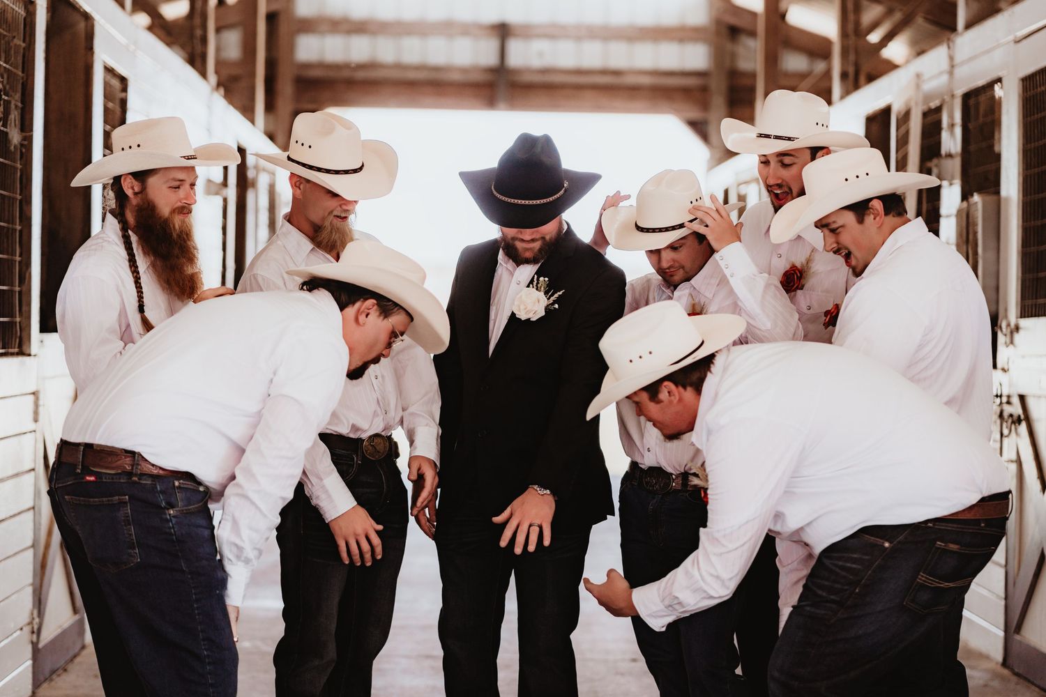 Wedding party huddles around groom in black suit and cowboy hat inside rustic barn venue.