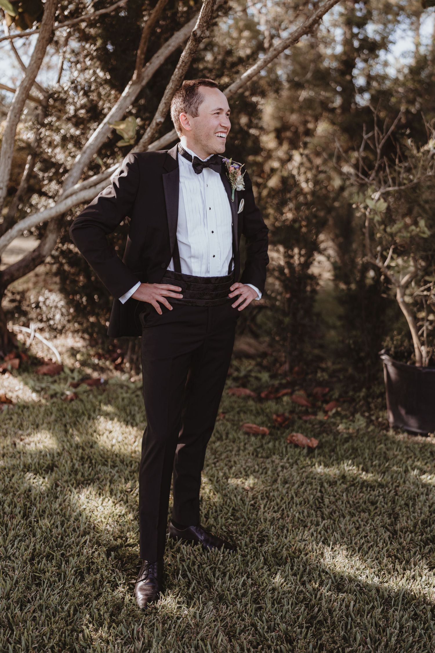 Handsome groom in black tuxedo with boutonniere smiling with hands on hips in a sunlit garden.