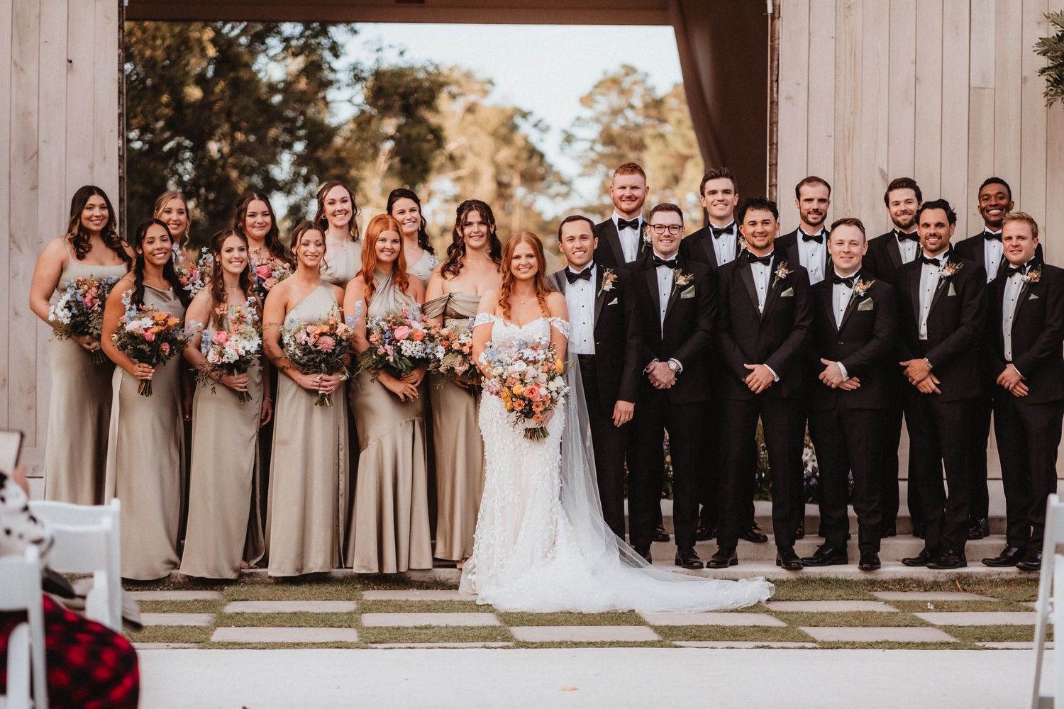 Large wedding party poses together, bridesmaids in champagne gowns and groomsmen in black tuxedos outside a barn venue.