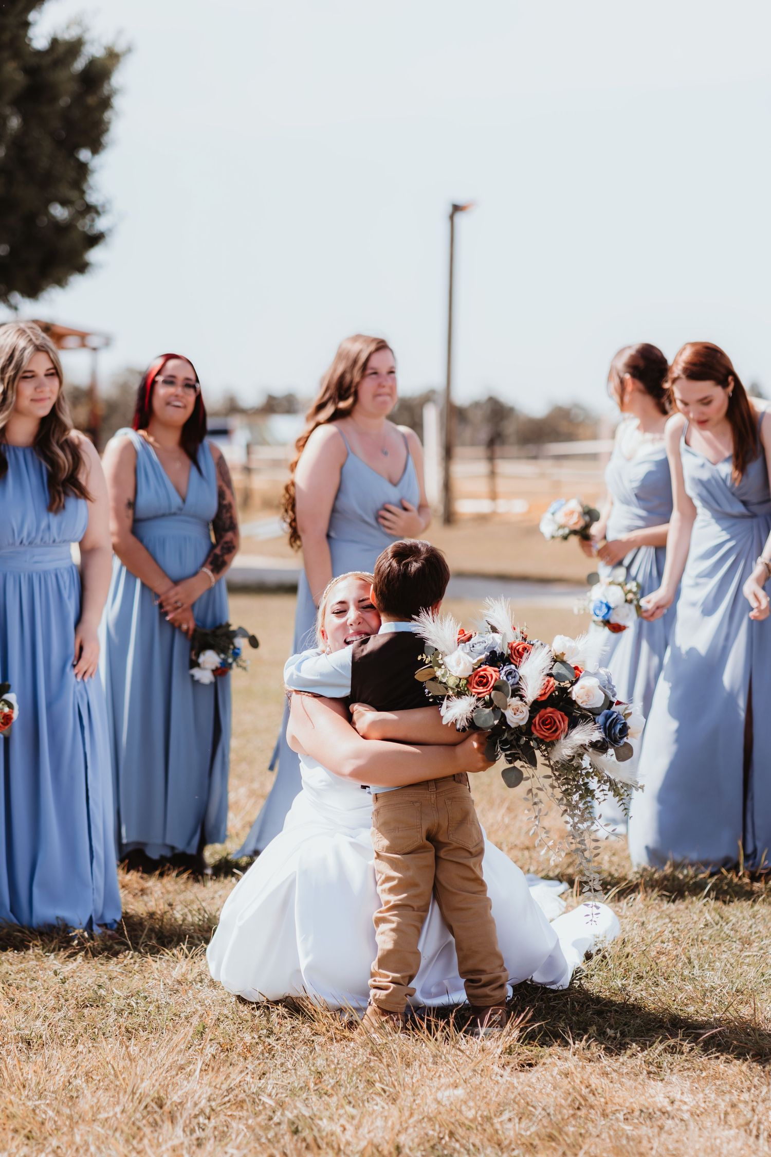 Bride in white wedding gown kneeling to hug a young boy while bridesmaids in blue dresses look on.