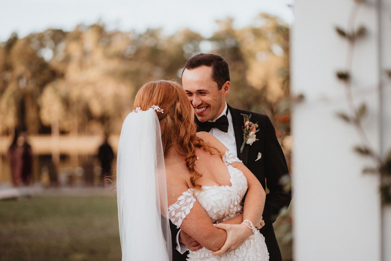 Groom in tuxedo laughs joyfully while embracing red-haired bride in floral lace wedding dress.