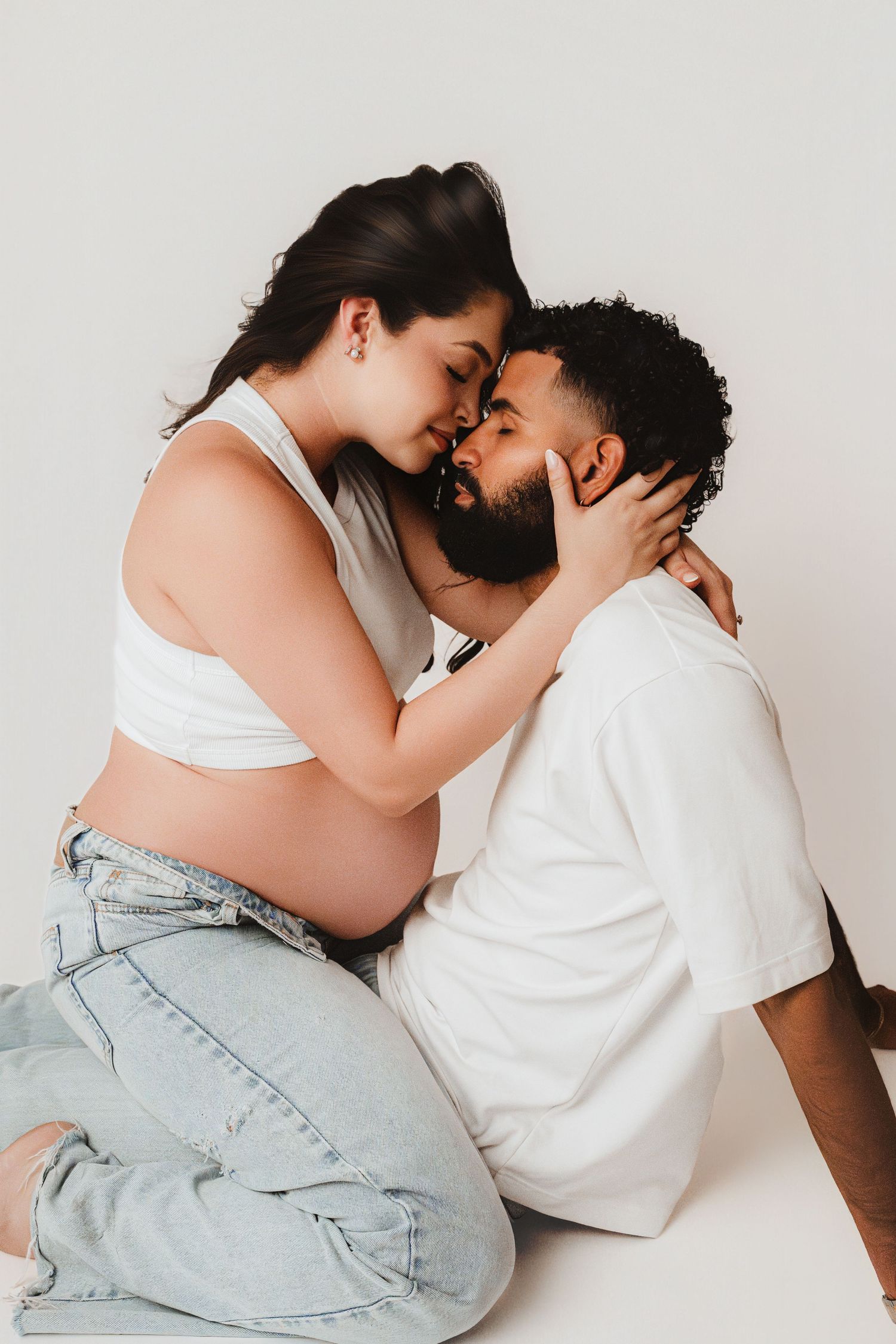 Pregnant couple sharing an intimate moment, touching foreheads while sitting on white studio floor.