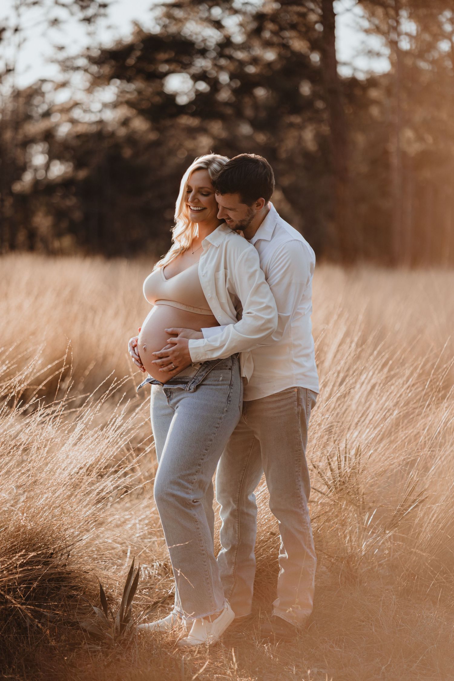 Happy pregnant couple embracing in warm sunlit field, man kissing woman's head.