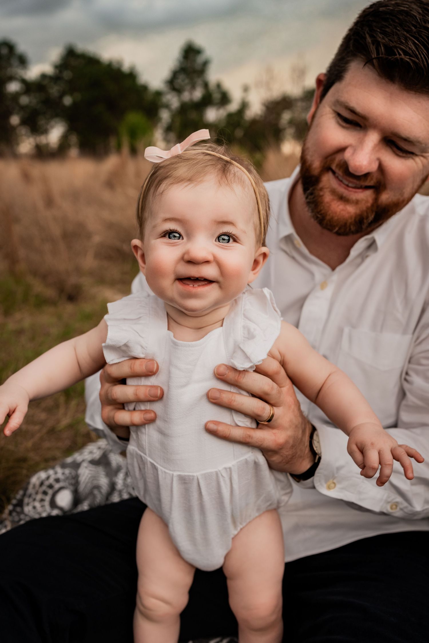 Bearded father in white shirt smiling at happy baby girl in white ruffle romper in a golden field.