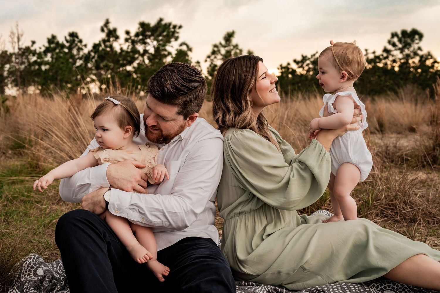 Parents sitting back to back on a patterned blanket each cuddling a baby in a warm sunset field.