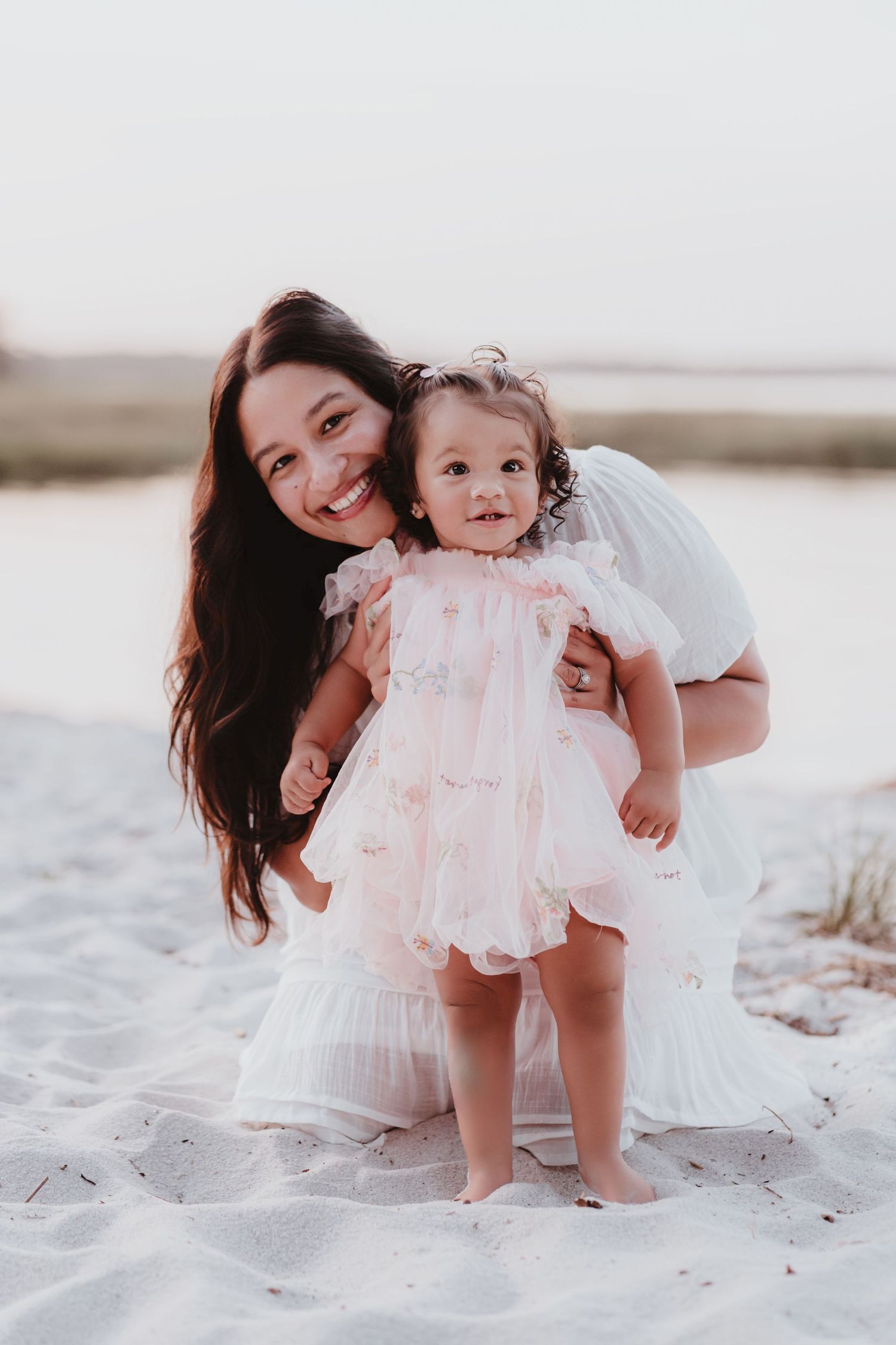 Happy mother kneeling behind standing toddler girl in pink dress on beach.