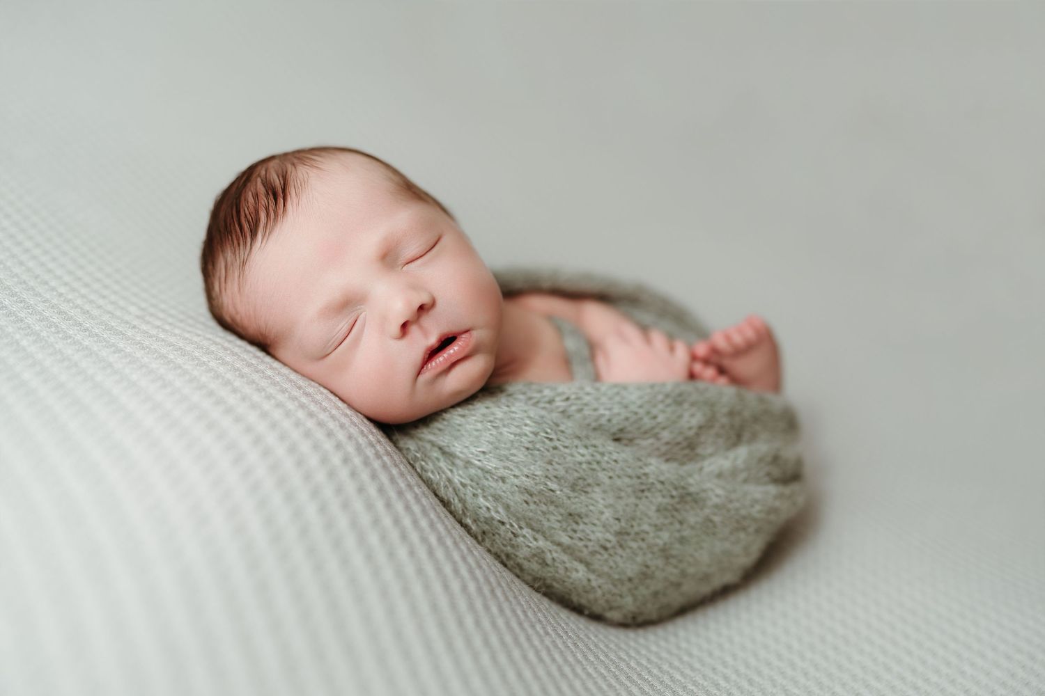Newborn baby swaddled in sage green wrap sleeping on gray textured blanket.