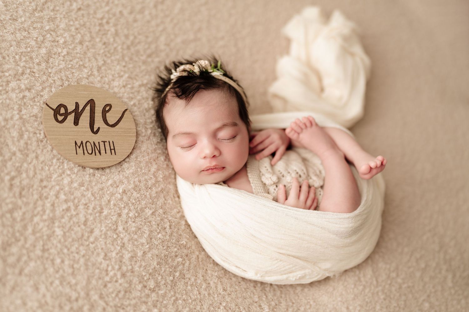 Sleeping newborn baby girl wrapped in cream swaddle beside a one month milestone wooden sign.