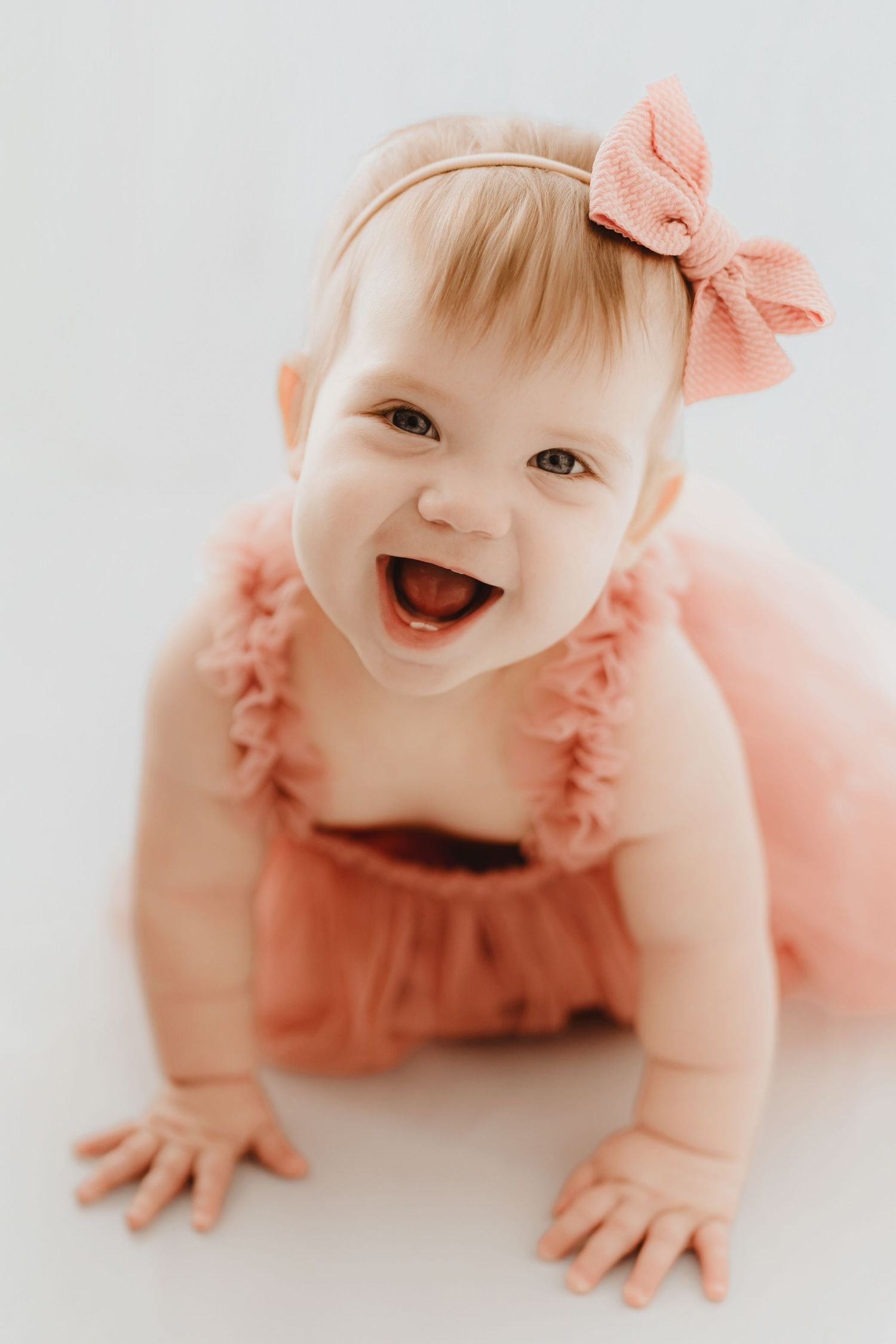 Laughing baby girl in coral ruffle romper and bow headband joyfully crawling on white studio floor.