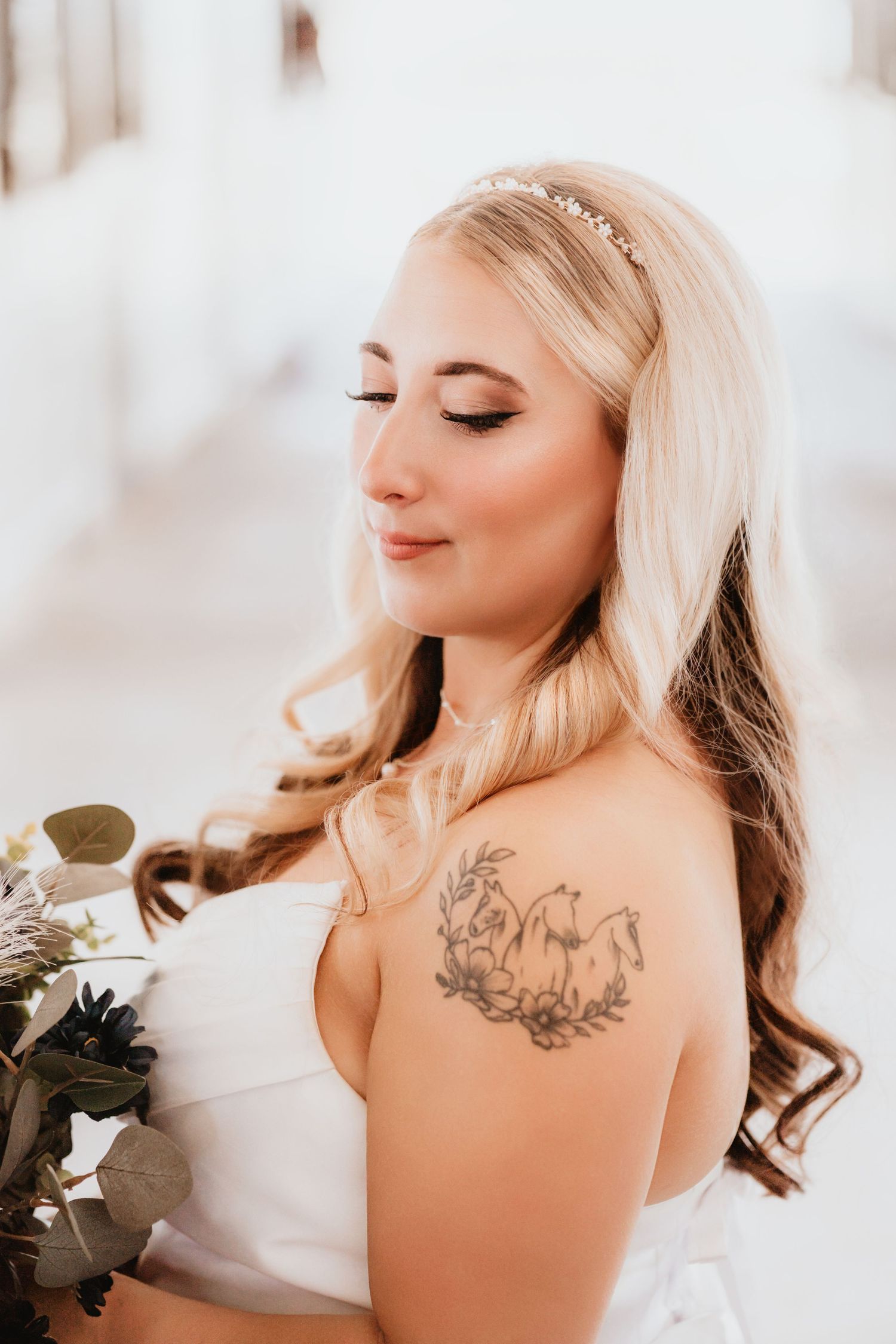 Close-up of a blonde bride with a horse tattoo on her shoulder, eyes closed, holding a floral bouquet.