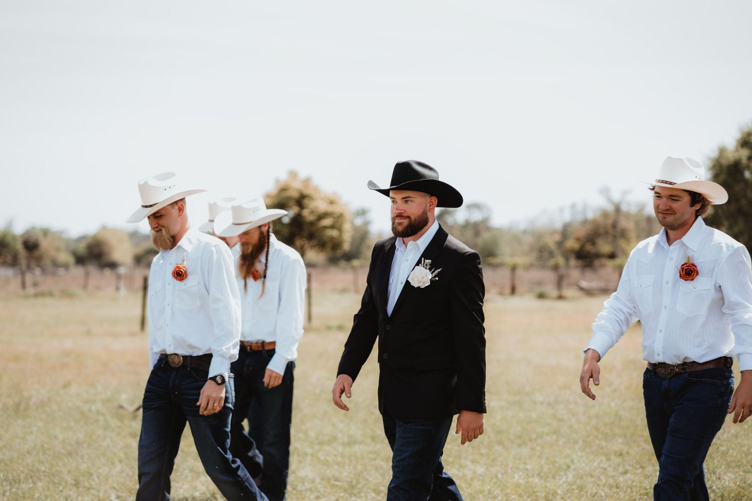 Groom in black cowboy hat and suit leads groomsmen in white shirts across a rustic ranch field.