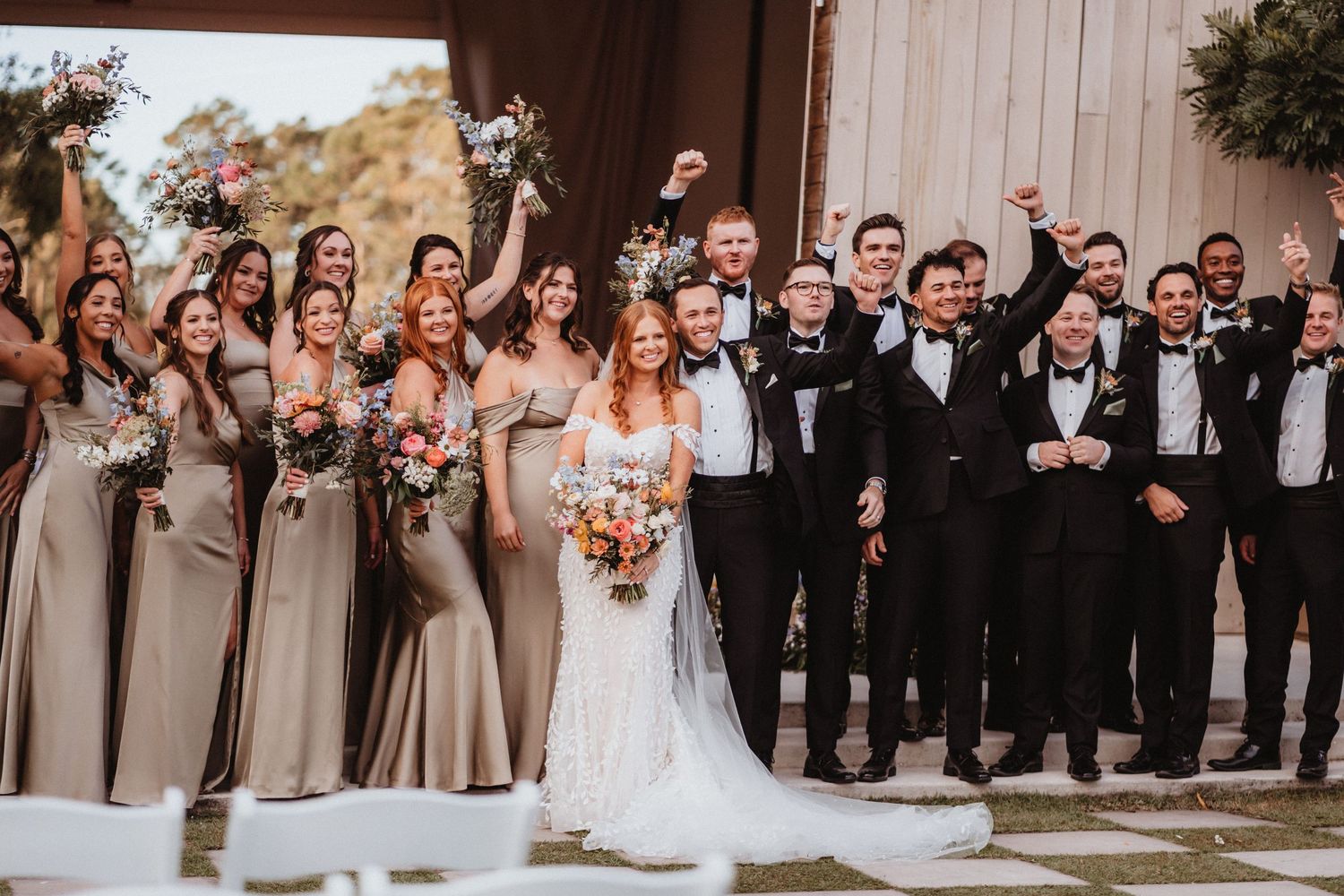 Large wedding party poses together outdoors, bridesmaids in taupe gowns and groomsmen in black tuxedos.