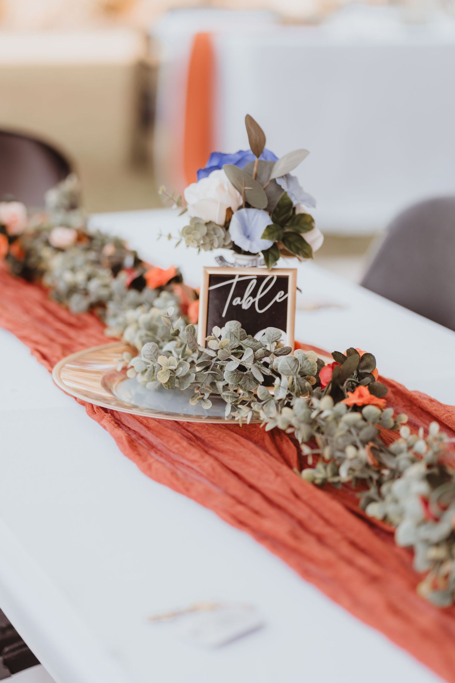 Rustic wedding table centerpiece with terracotta runner, eucalyptus garland, and small chalkboard table number sign.
