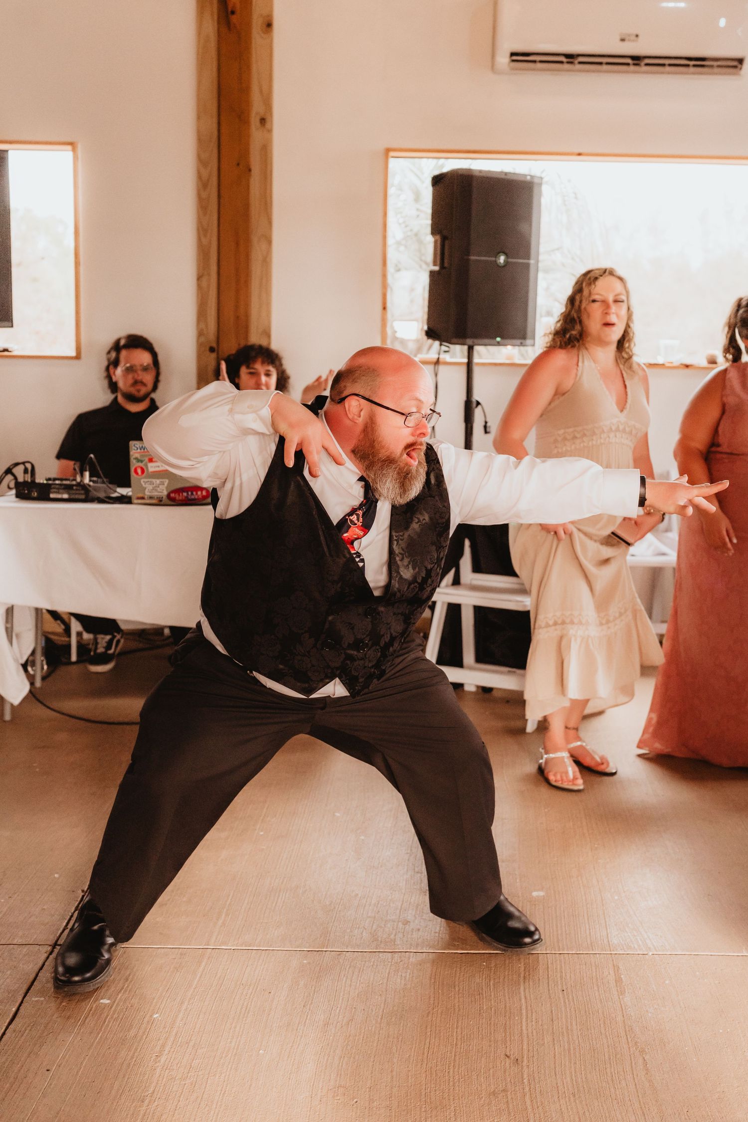 Bald man in vest performs energetic dance moves at wedding reception on dance floor.
