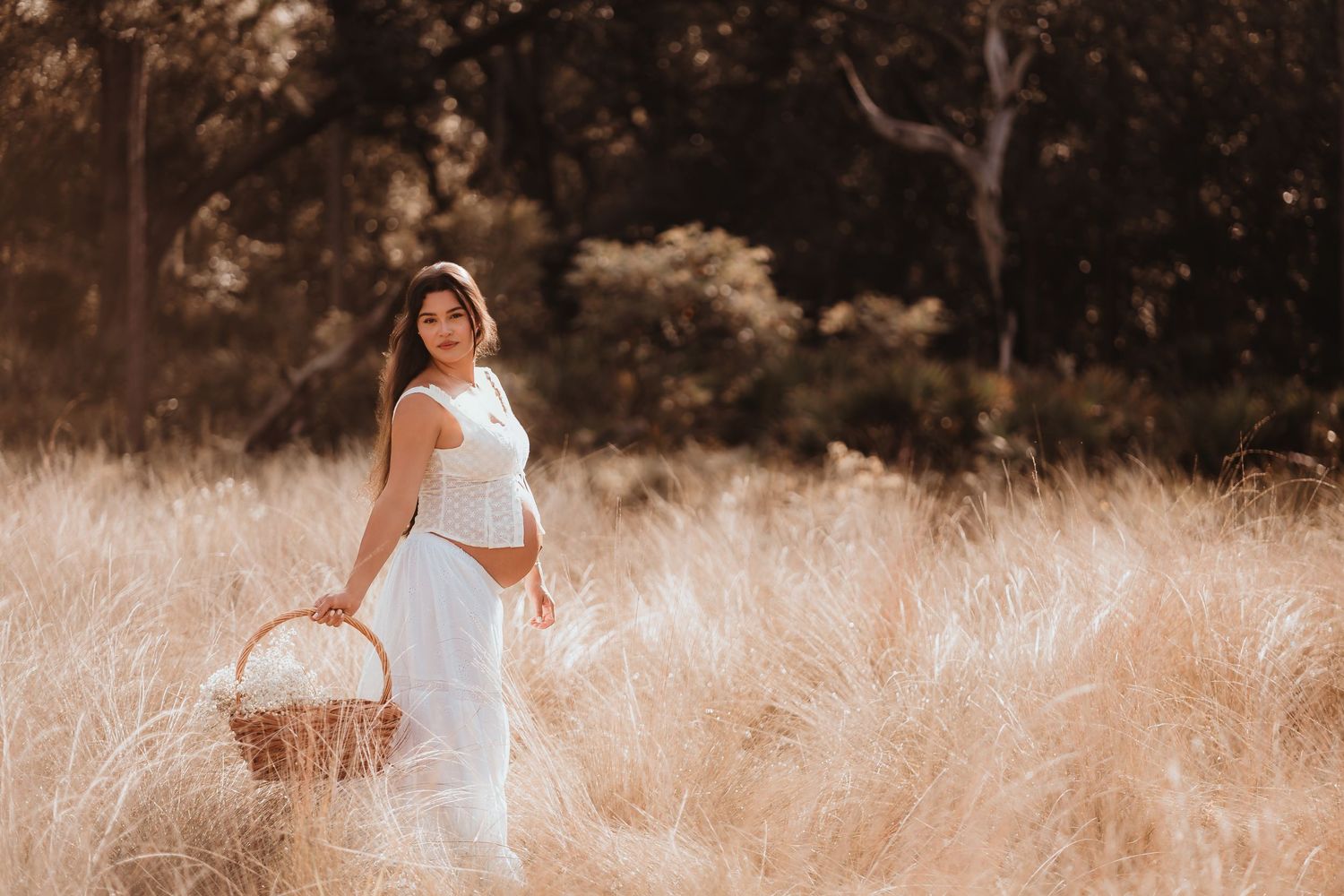 Pregnant woman in white dress holding wicker basket in sun-drenched golden grass field.