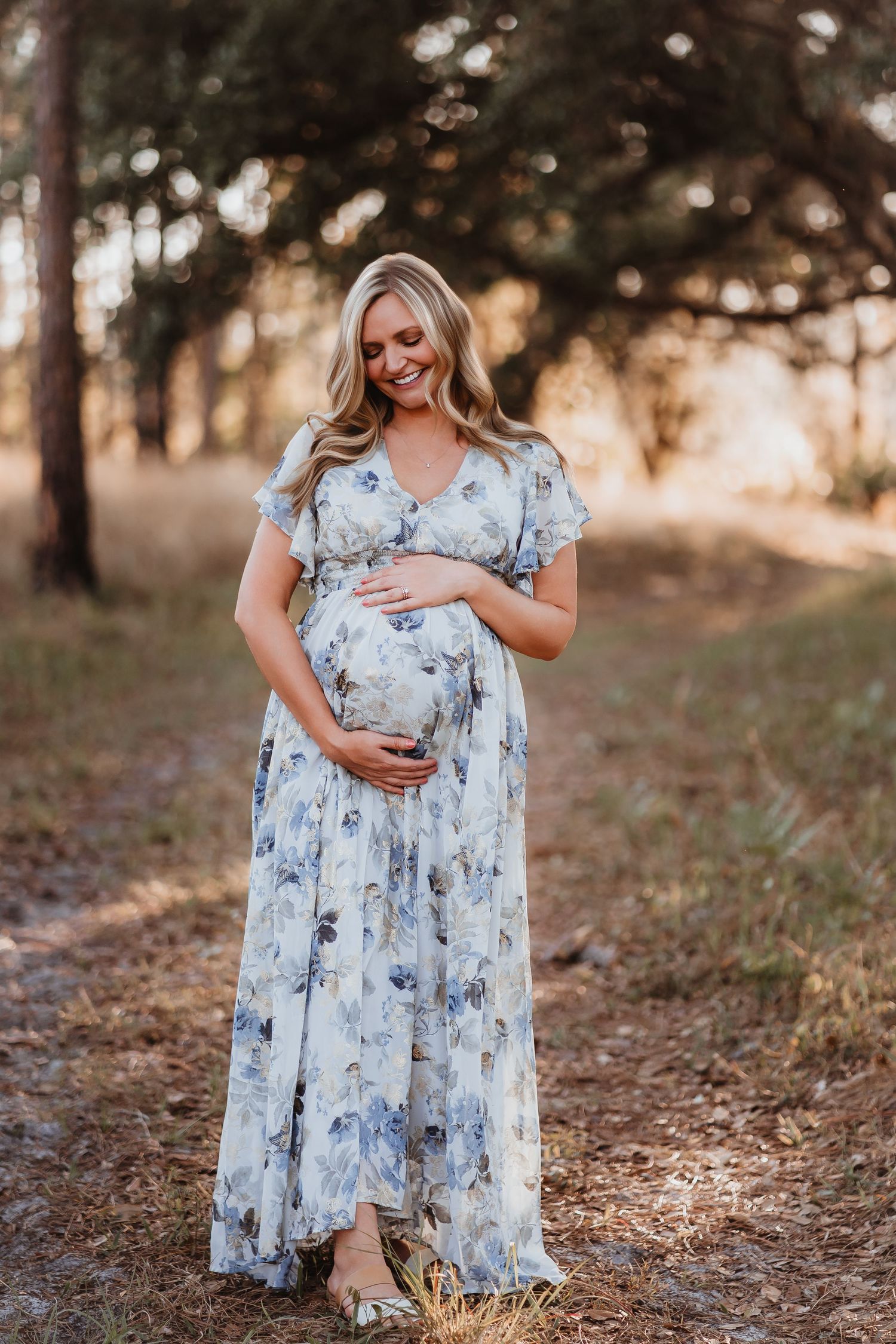 Smiling blonde pregnant woman in floral maxi dress cradling baby bump in forest setting.