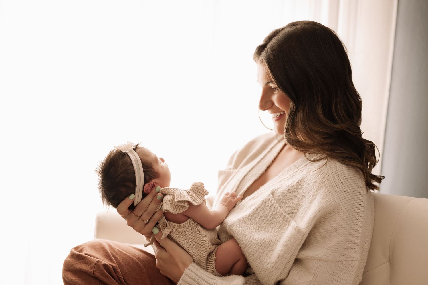 Smiling brunette mother in cream sweater tenderly holding newborn baby by bright window.