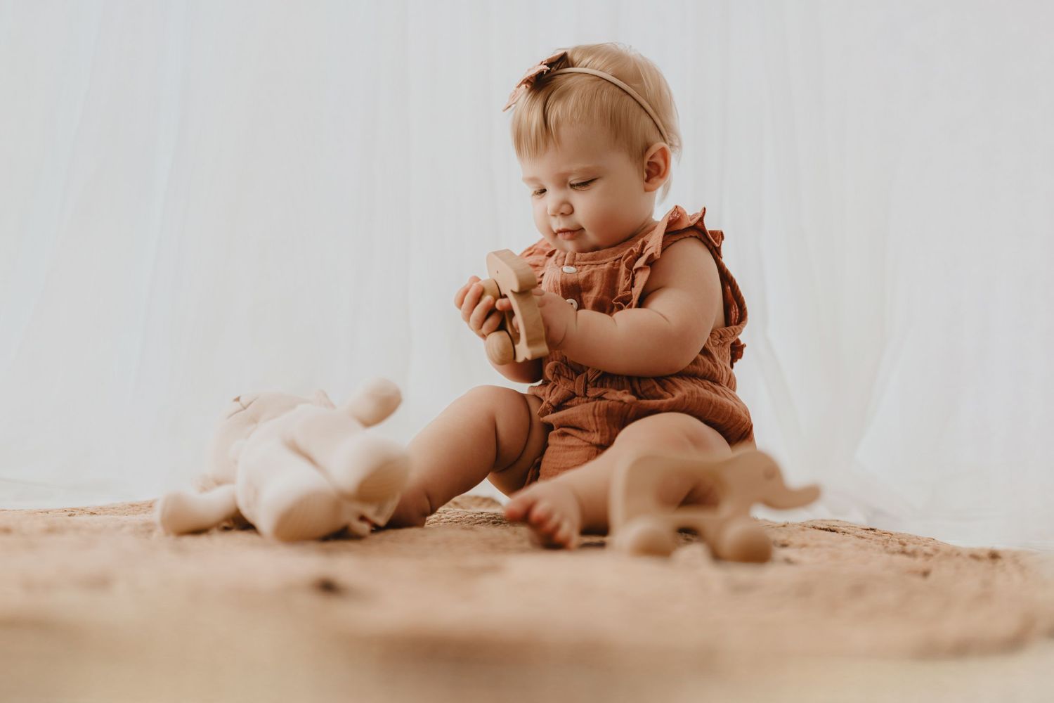 Baby girl in rust romper sits on jute rug playing with wooden toys and stuffed bunny.