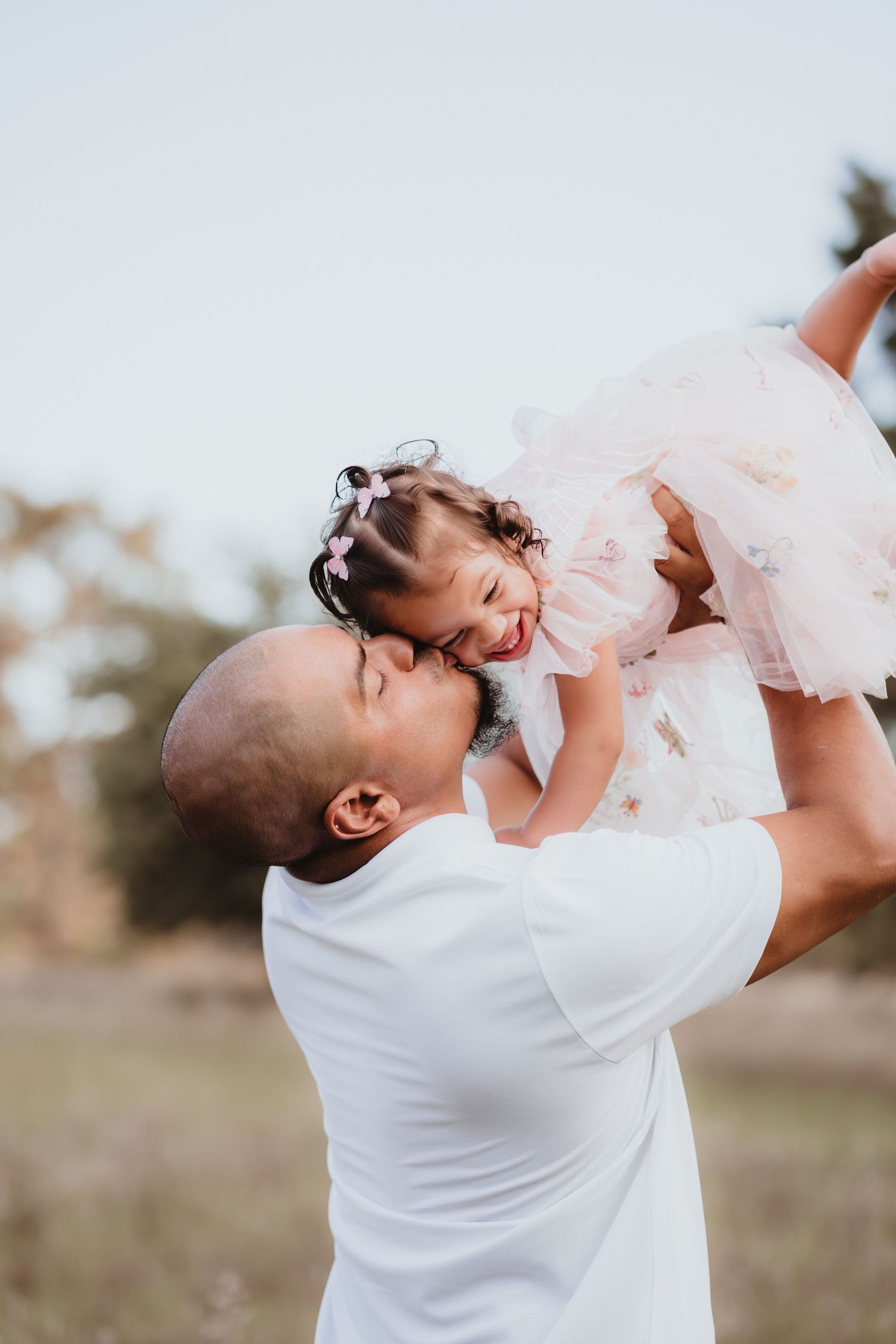 Tender moment as father lifts and kisses his smiling toddler daughter in a pink tulle dress.