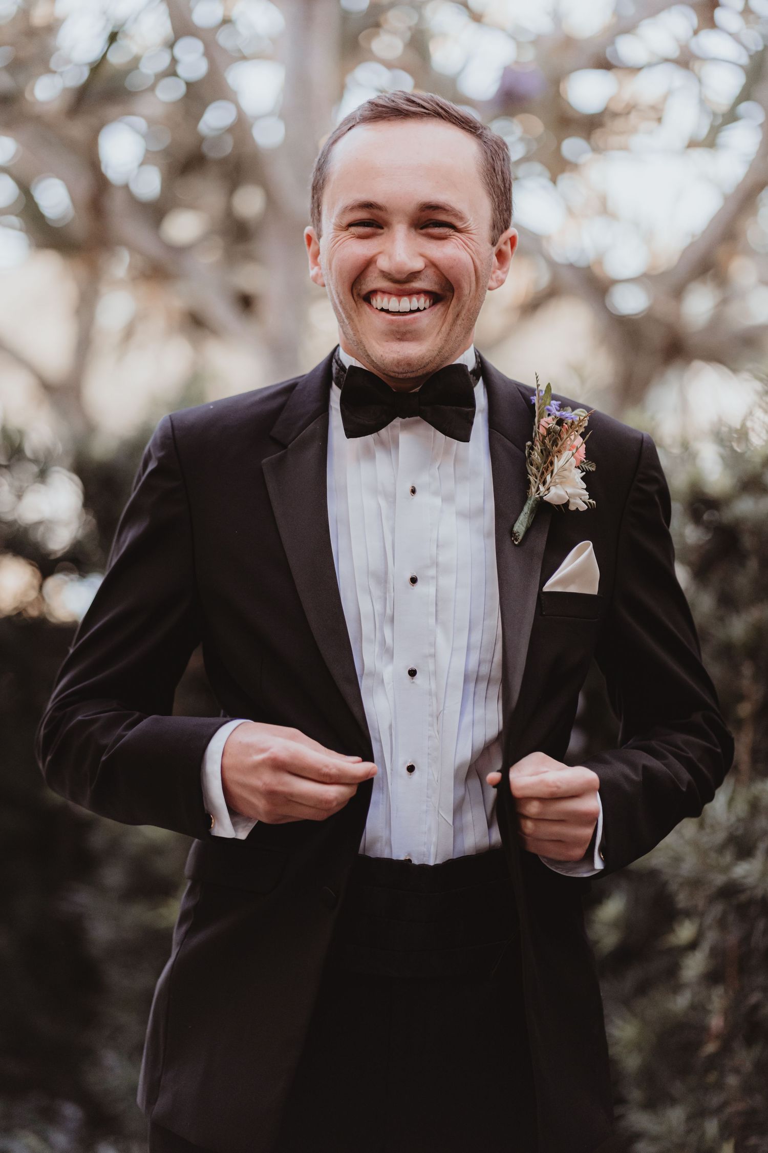 Cheerful groom in black tuxedo adjusting his jacket while smiling outdoors.