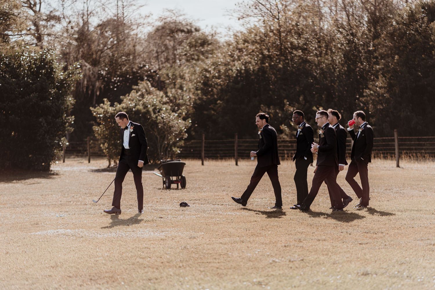 Groomsmen in dark suits walk together across a sunny open field during wedding portraits.