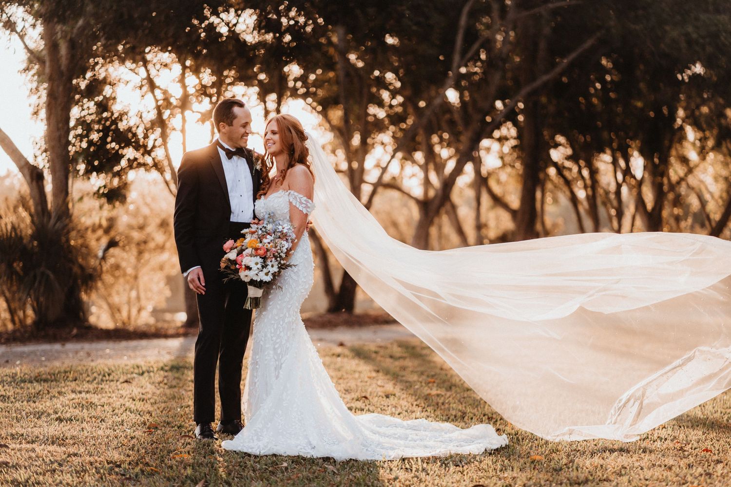 Bride and groom embrace at sunset under oak trees, her cathedral veil dramatically flowing in the wind.