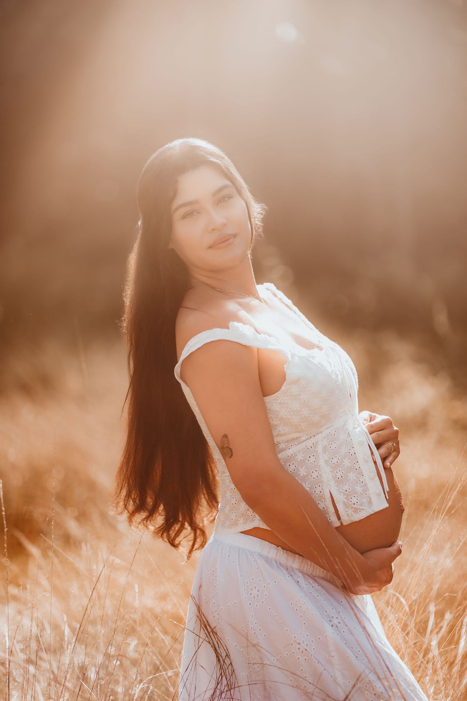 Beautiful pregnant woman with long dark hair in white dress posing in glowing golden field at sunset.