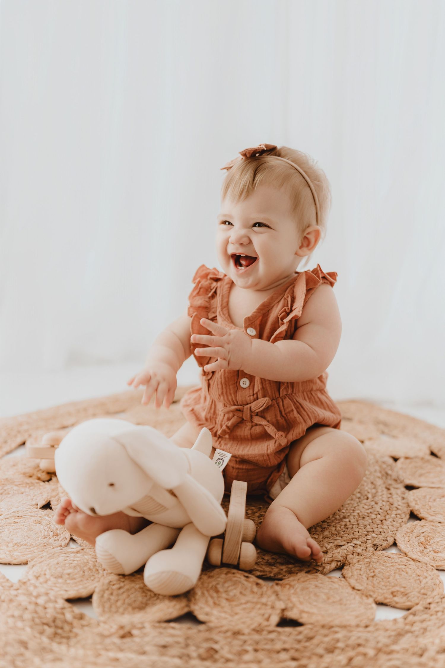Giggling baby girl in rust ruffle romper playing with stuffed bunny toy on round woven rug.