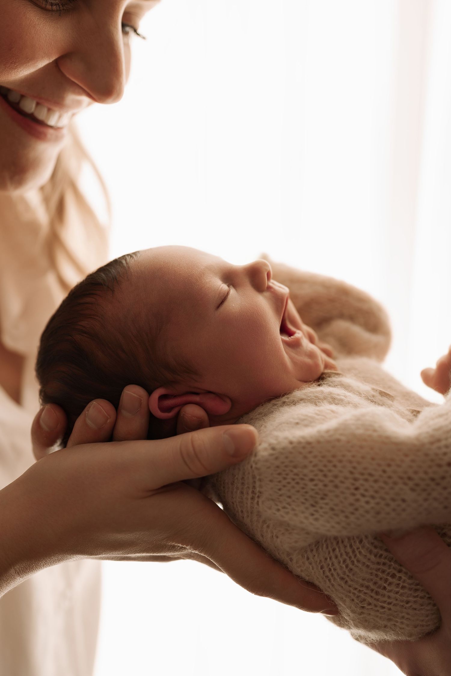 Smiling mother cradling yawning newborn baby wrapped in soft cream knit blanket.
