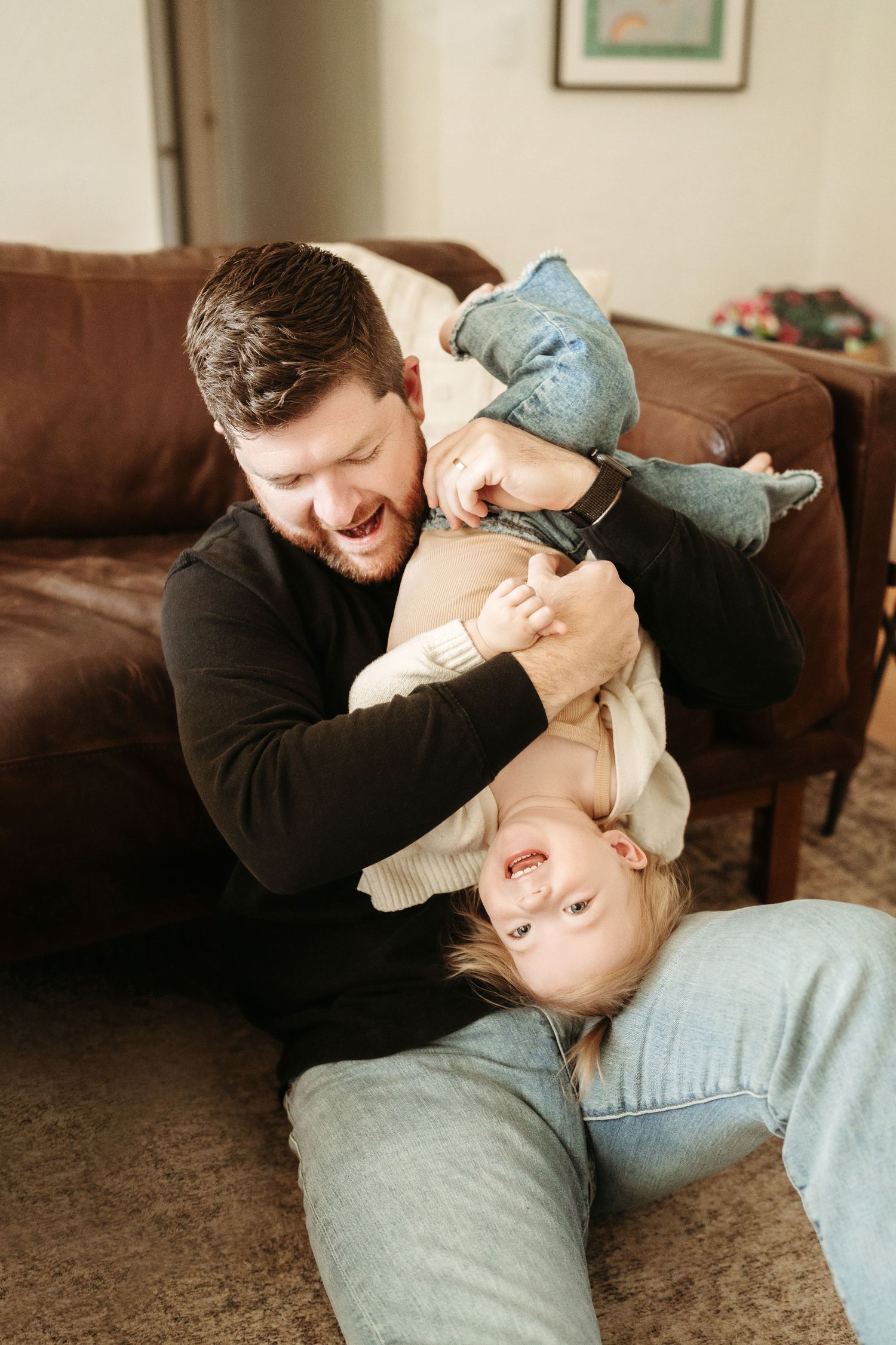 Father playfully holding toddler upside down while both laugh together in a cozy living room.