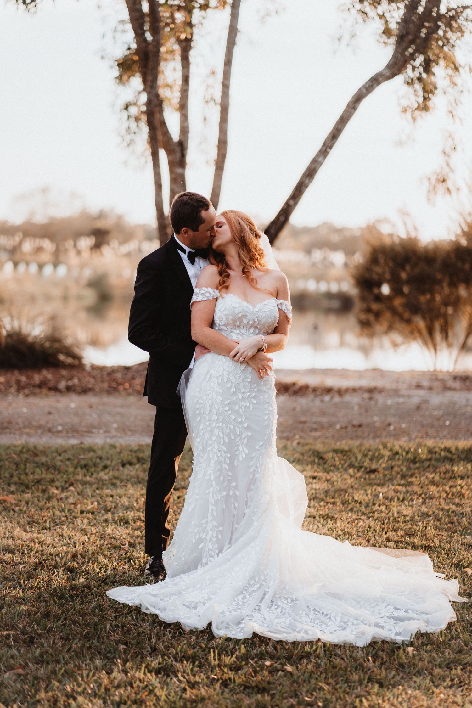 Bride and groom share a kiss outdoors at sunset near a lake, bride wearing off-shoulder lace wedding gown.