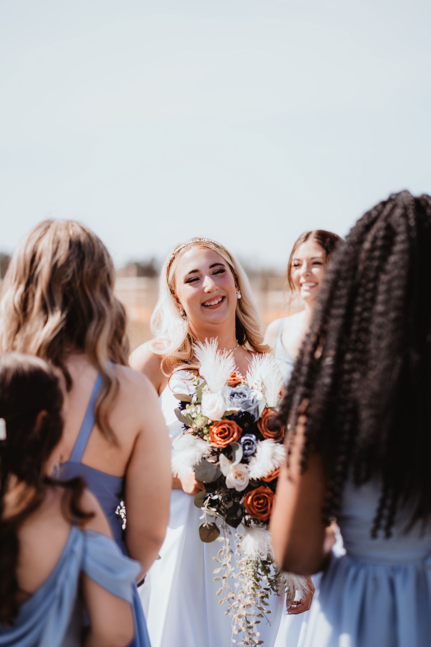 Smiling bride in white dress holds colorful bouquet surrounded by bridesmaids in light blue dresses outdoors.
