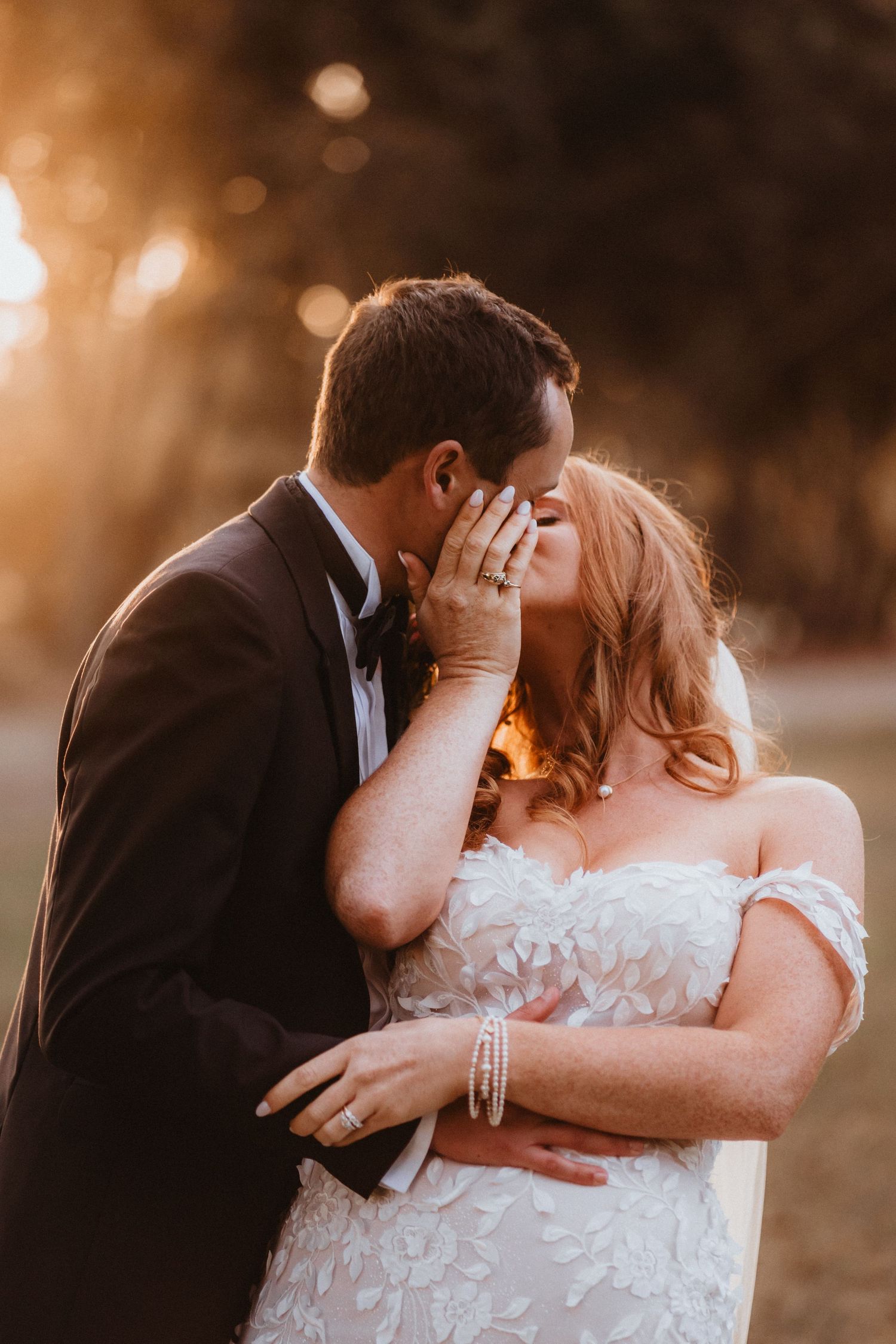 Couple shares a romantic golden hour kiss, bride in lace off-shoulder gown with pearl bracelet and ring visible.
