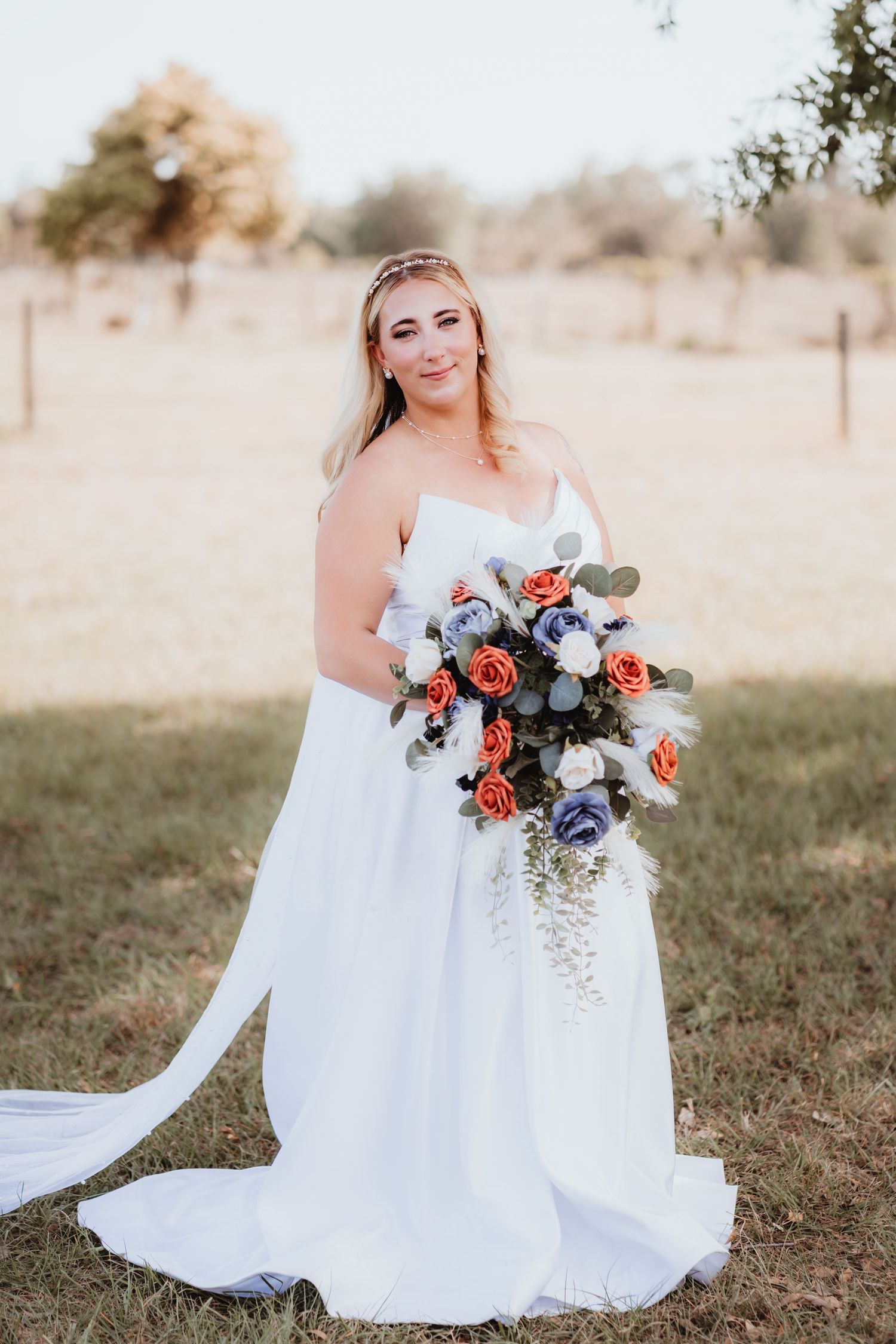 Blonde bride in strapless white gown with long train holding vibrant orange and blue rose bouquet.