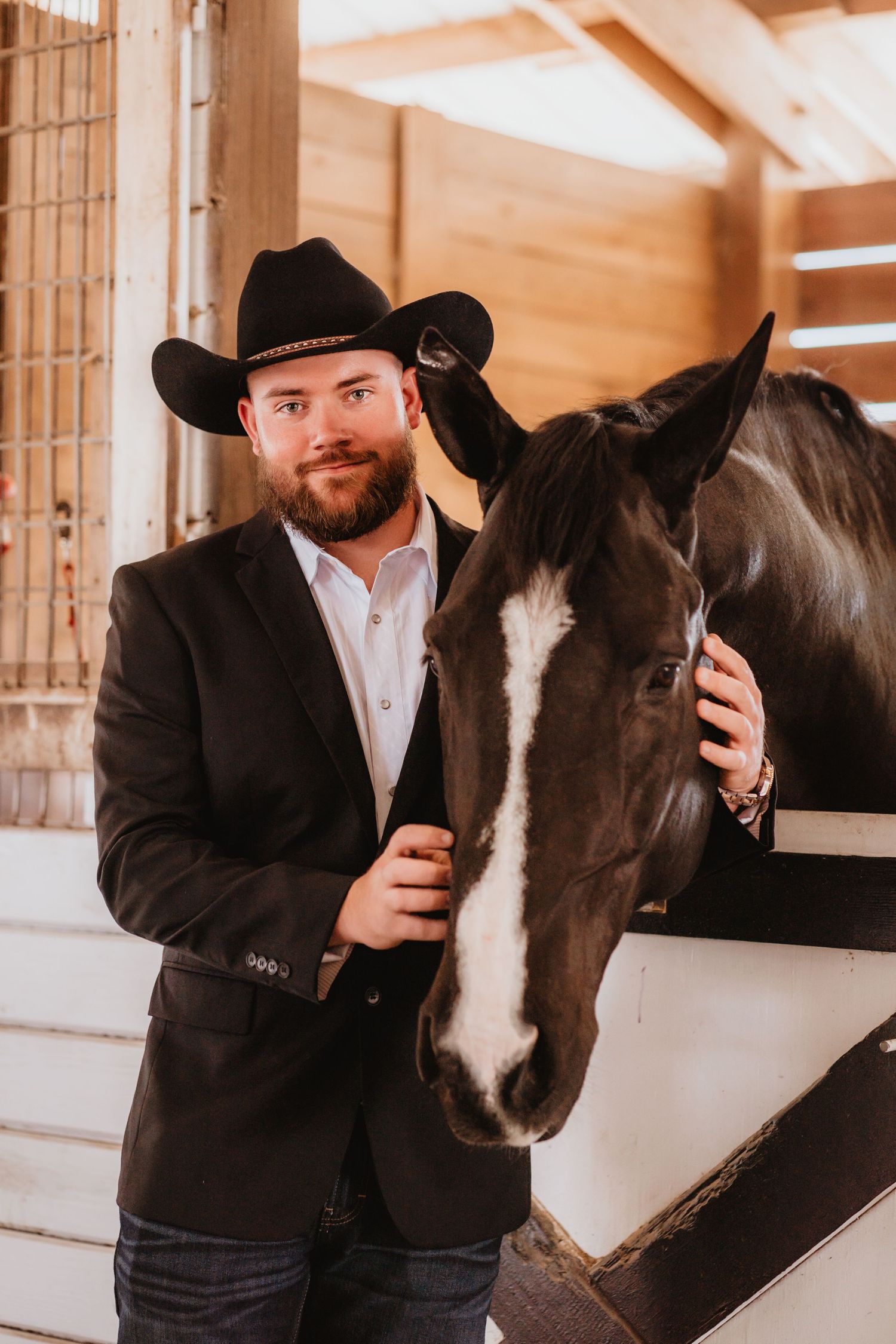 Handsome groom in black cowboy hat and blazer poses with dark horse inside rustic stable.