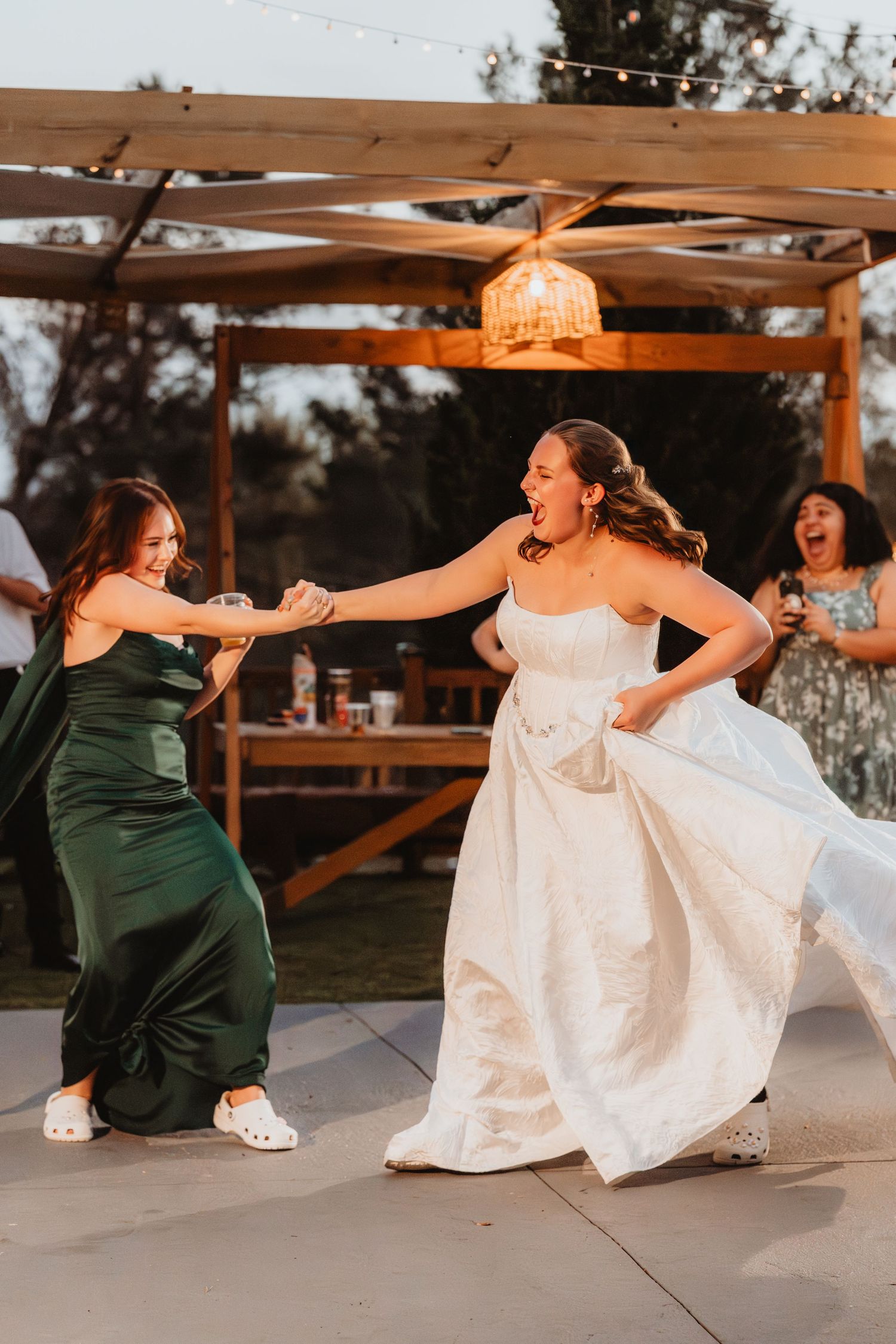 Bride in white gown dancing joyfully with bridesmaid in green dress at outdoor reception.