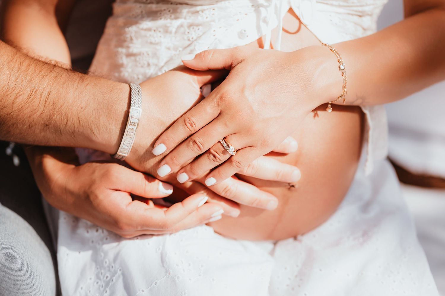 Close-up of couple's hands gently resting on pregnant woman's belly in white dress.