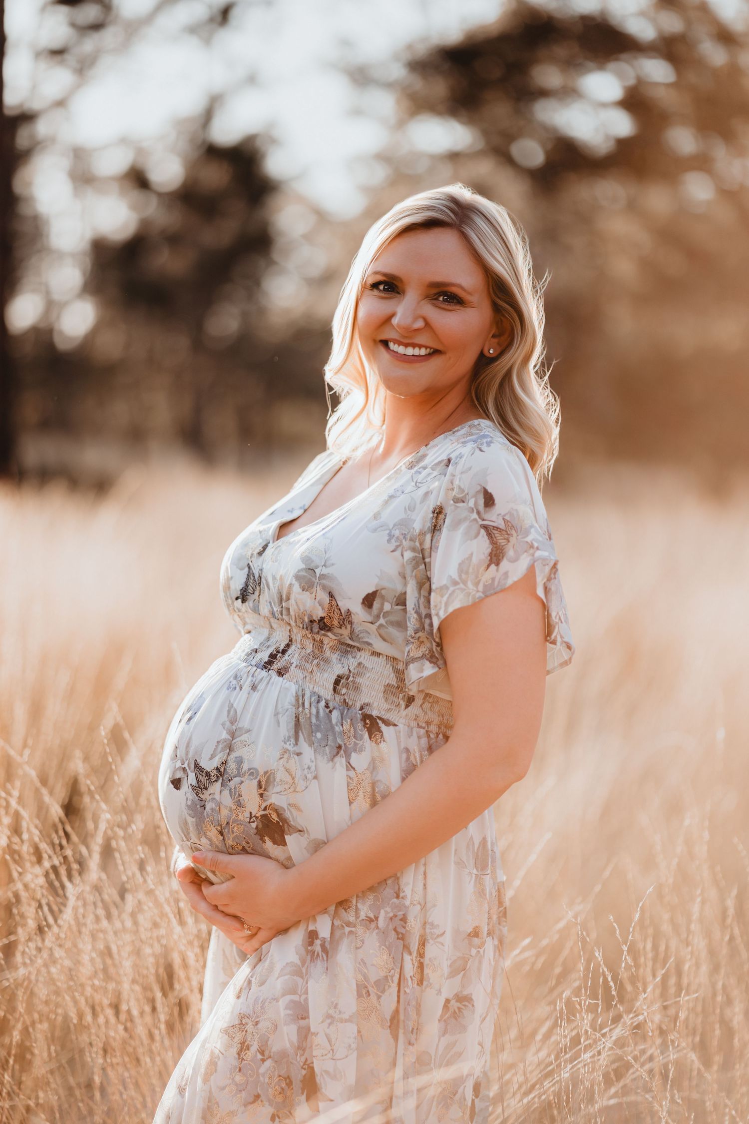 Smiling pregnant woman in floral dress holds baby bump in warm sunlit field during maternity photoshoot.