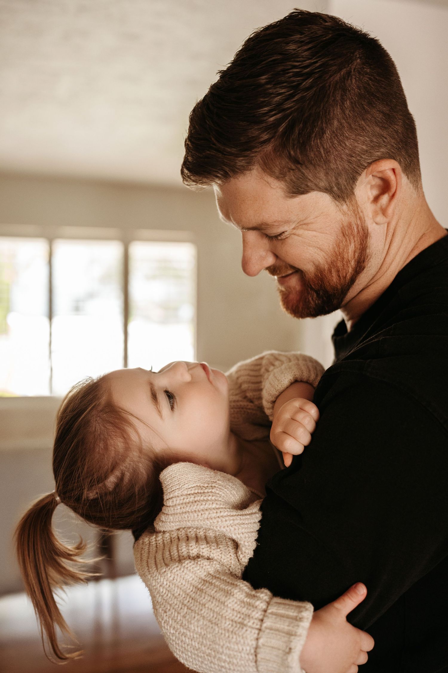 A bearded father smiles warmly while holding his young daughter close in a bright home.